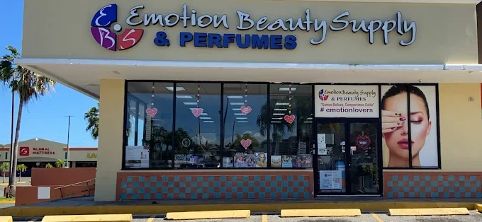Exterior of a store called 'Emotion Beauty Supply & Perfumes' with large glass windows decorated with pink heart-shaped ornaments. There is a promotional sign on the window and a large poster of a woman on the right side. The store is located in a shopping plaza with a Palm tree visible on the left.