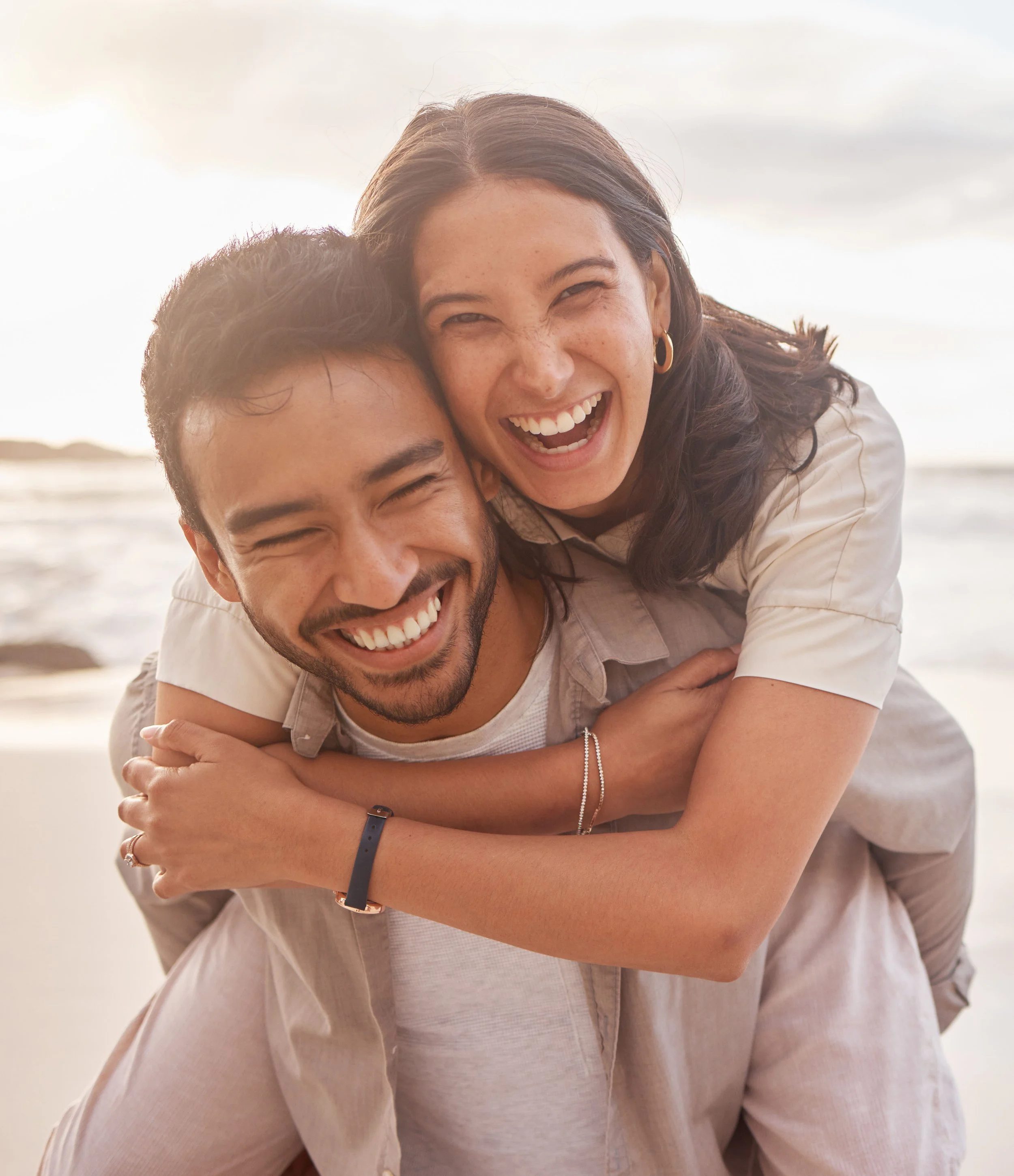 Close-up of a smiling woman with brown hair and gold earrings, hugging a man at the beach during sunset.