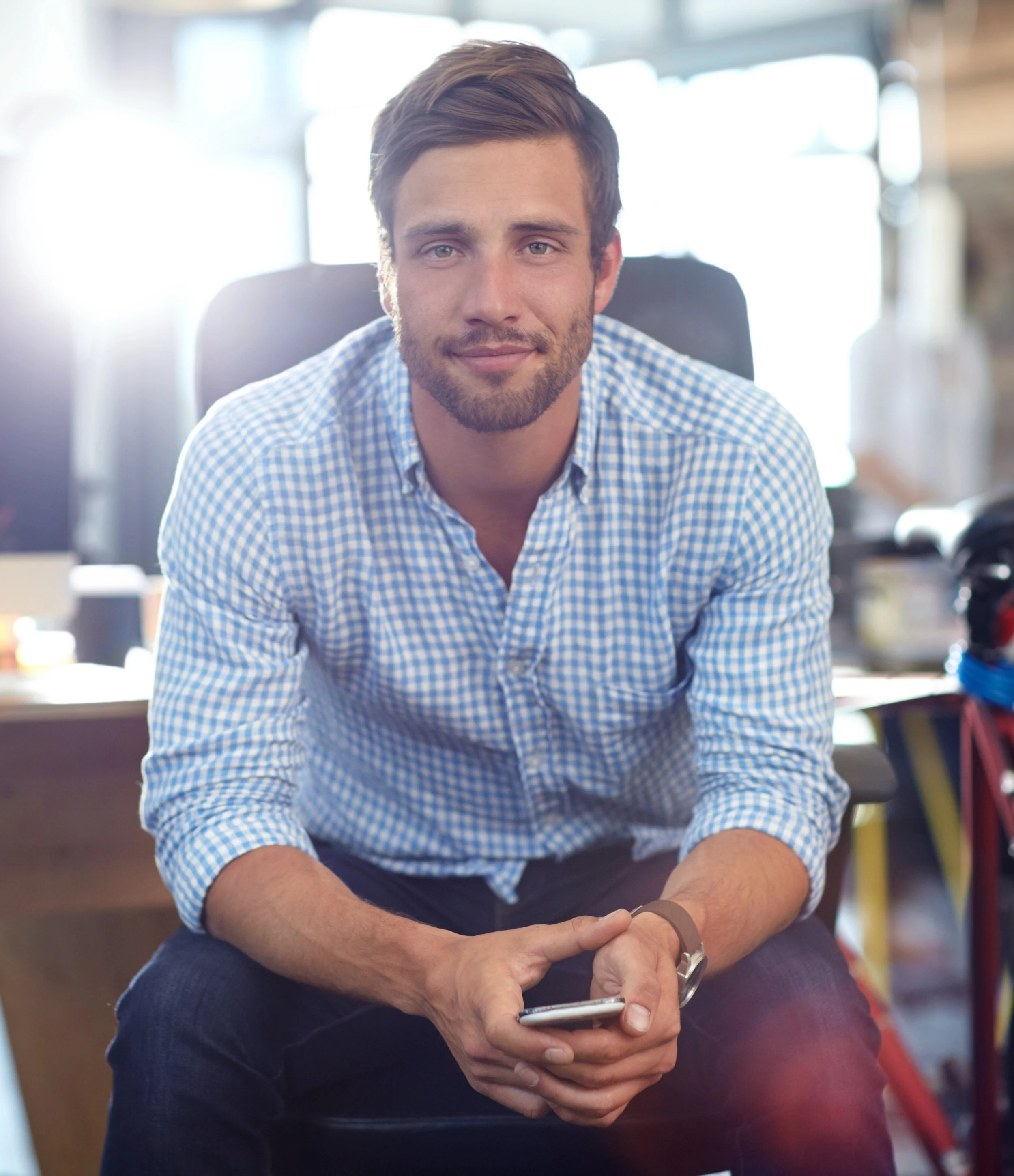 A young man with short brown hair and a beard, wearing a blue checkered shirt, sitting in an indoor space with bright natural light, looking at the camera, holding a smartphone in his hands.