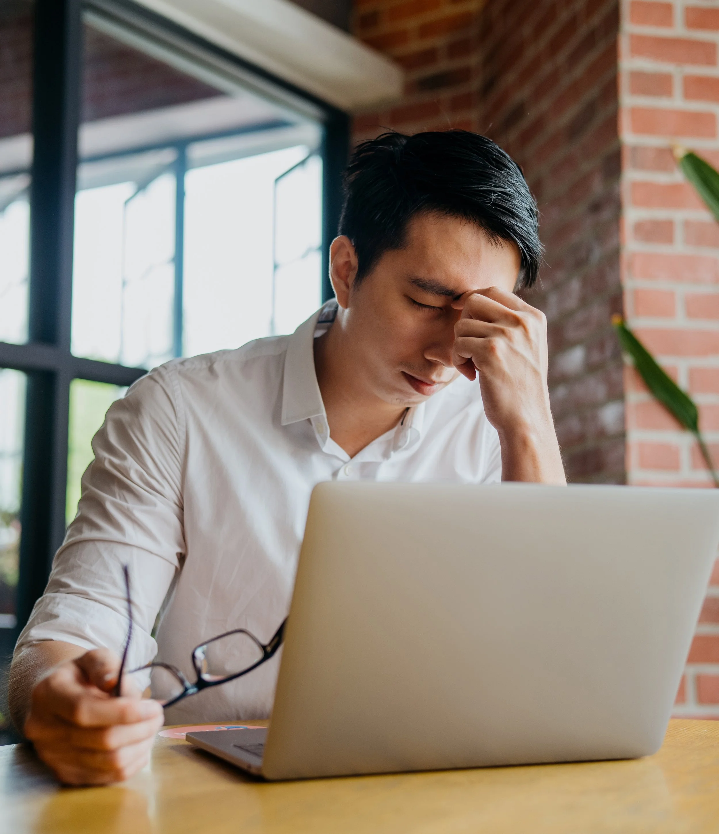A young man with black hair seated at a table, holding a pair of eyeglasses in his right hand, with a laptop in front of him, and appearing to be stressed or thinking, in a room with large windows and brick wall.