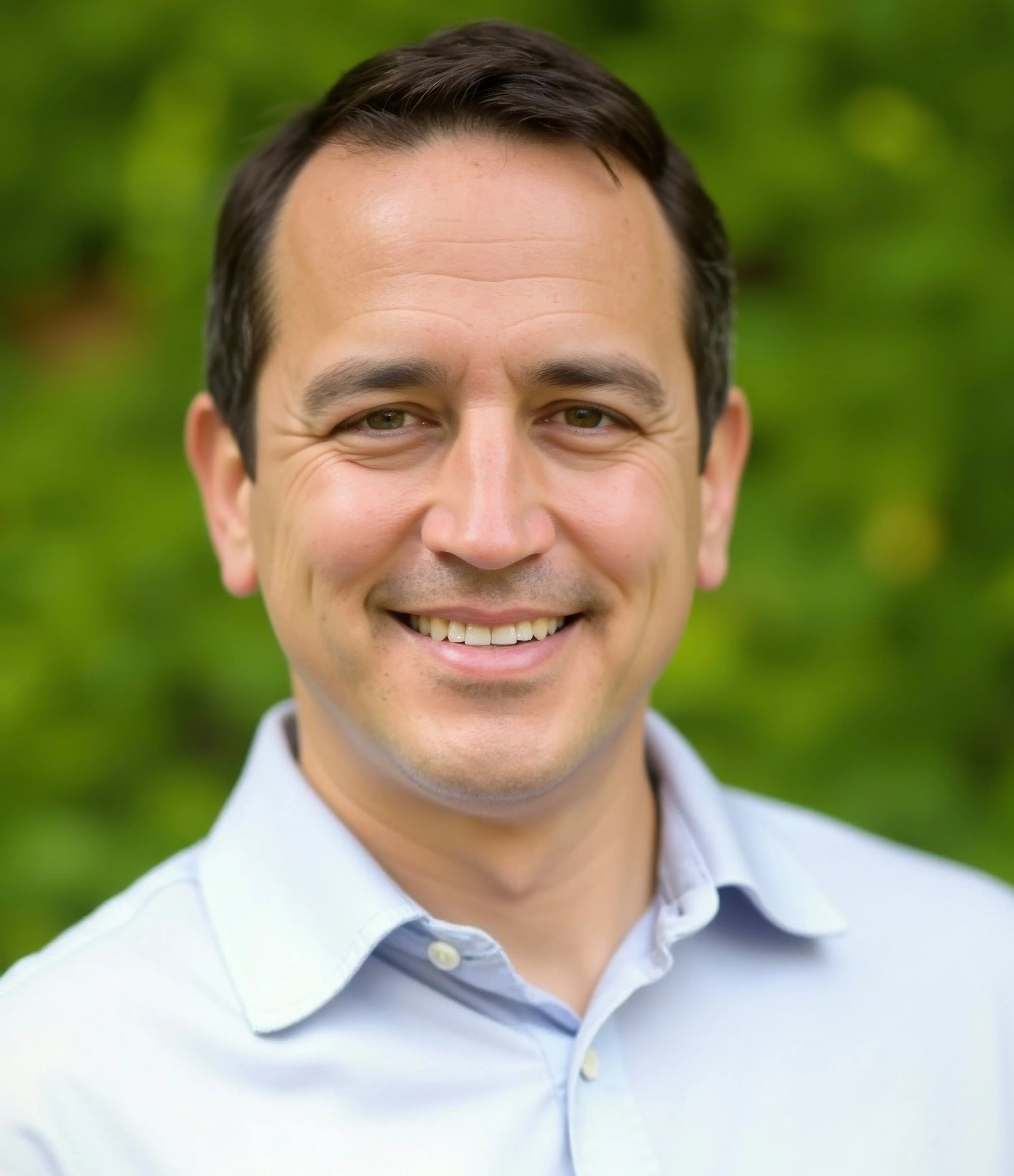 Close-up of a smiling man wearing a light blue collared shirt, with a blurred green outdoor background.