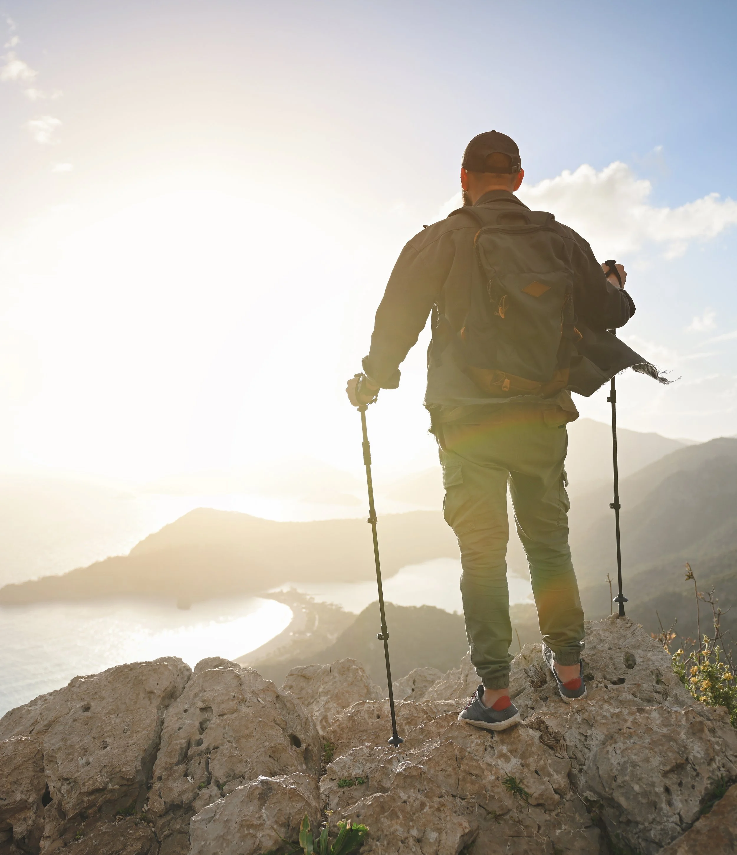 Man standing on a rocky ledge looking at a mountainous landscape with water, wearing hiking gear including a backpack, cap, and holding hiking poles, during sunset or sunrise.