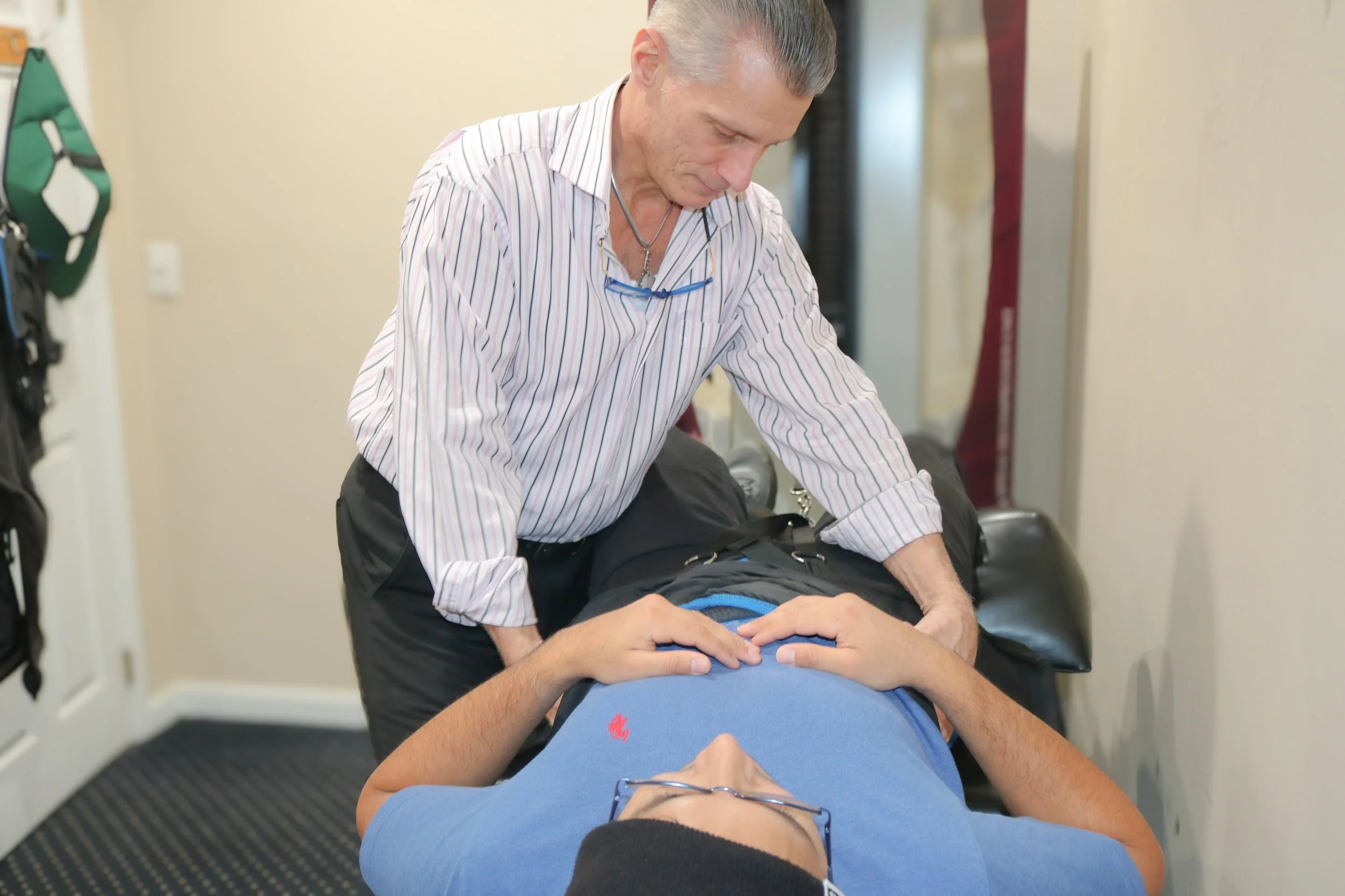 A chiropractor professional performing chest compressions on a person lying on a medical examination table. The person being treated is wearing glasses and a blue shirt.
