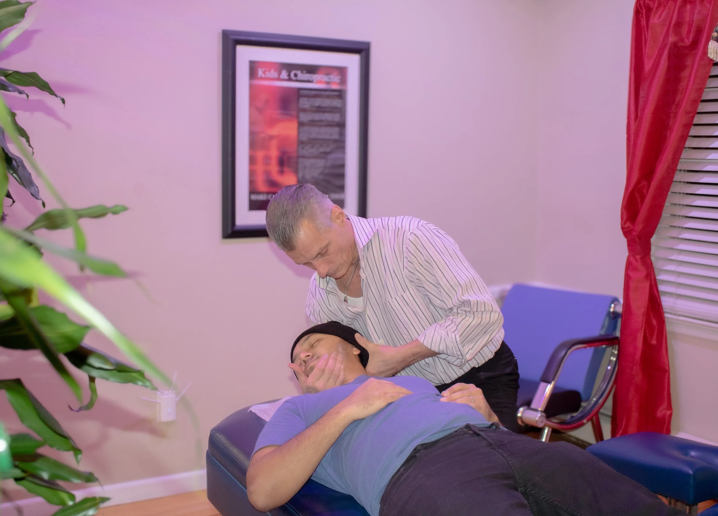 A person lying on a medical examination table having their neck examined by a chiropractor professional in an indoor setting, with a framed poster on the wall and a window with red curtains.