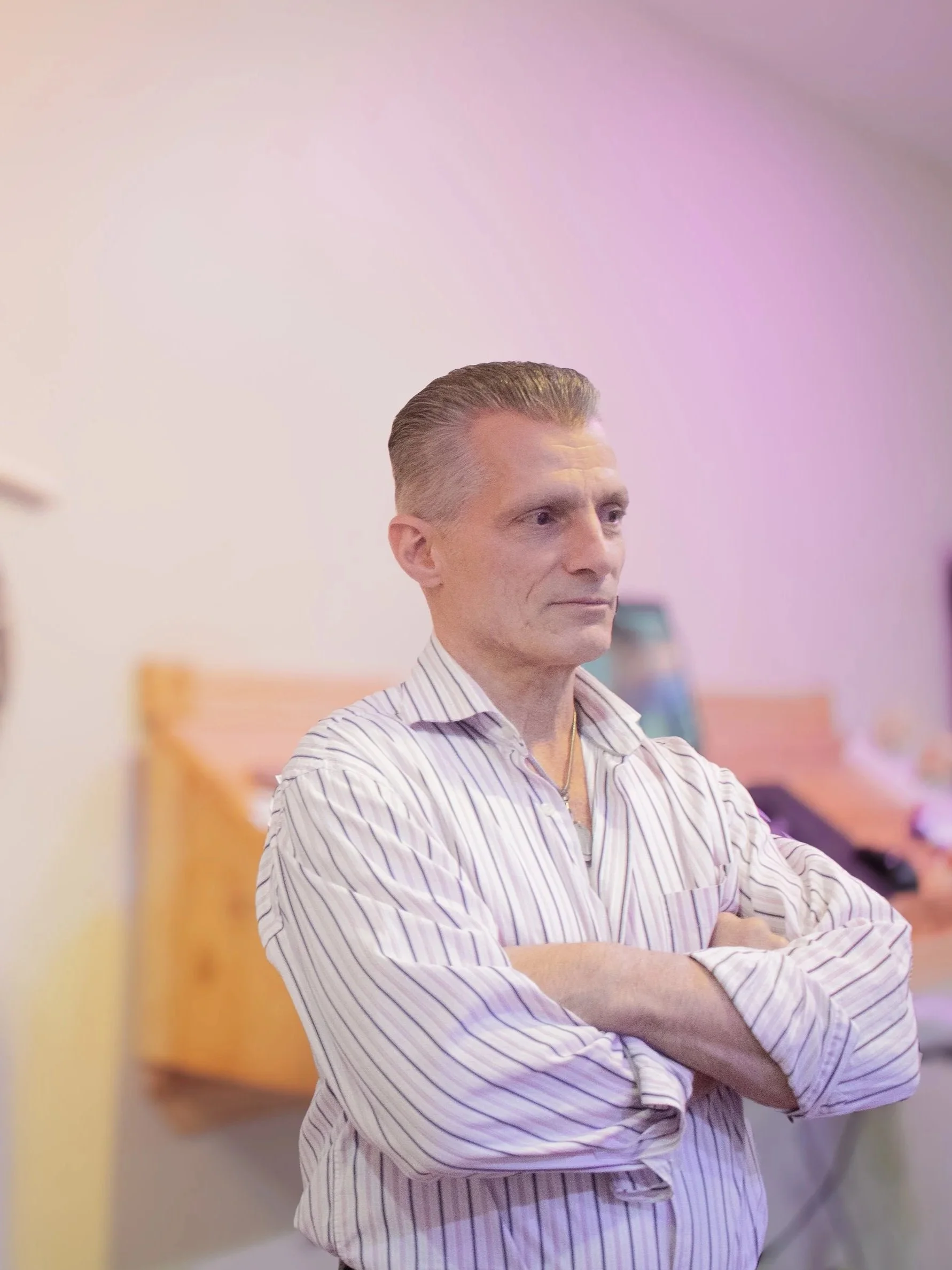 A chiropractor professional with slicked-back hair, wearing a striped dress shirt, is standing with his arms crossed in a room with soft pink lighting, wooden furniture, and blurred objects in the background.