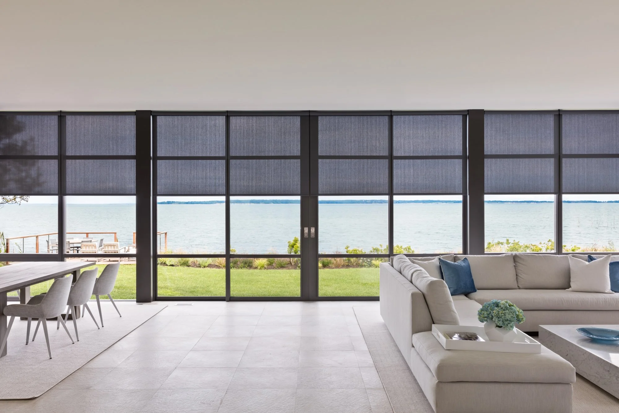 Living room with large glass windows overlooking water, patio with chairs outside, white sofa with blue pillows, dining chairs, and a vase with flowers on a coffee table.