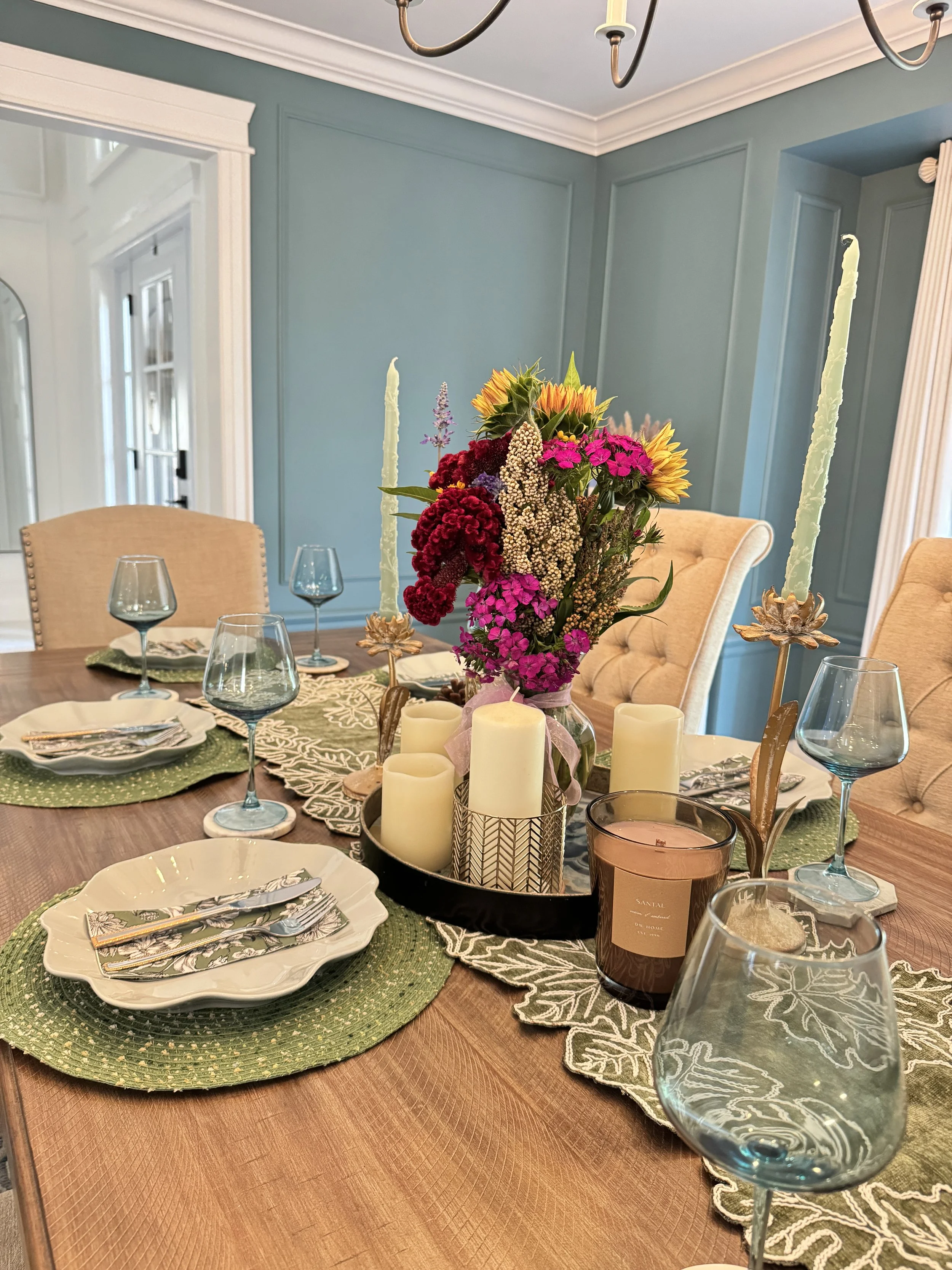 A dining table decorated with a bouquet of colorful flowers, candles, glassware, and plates in a dining room with light blue walls.
