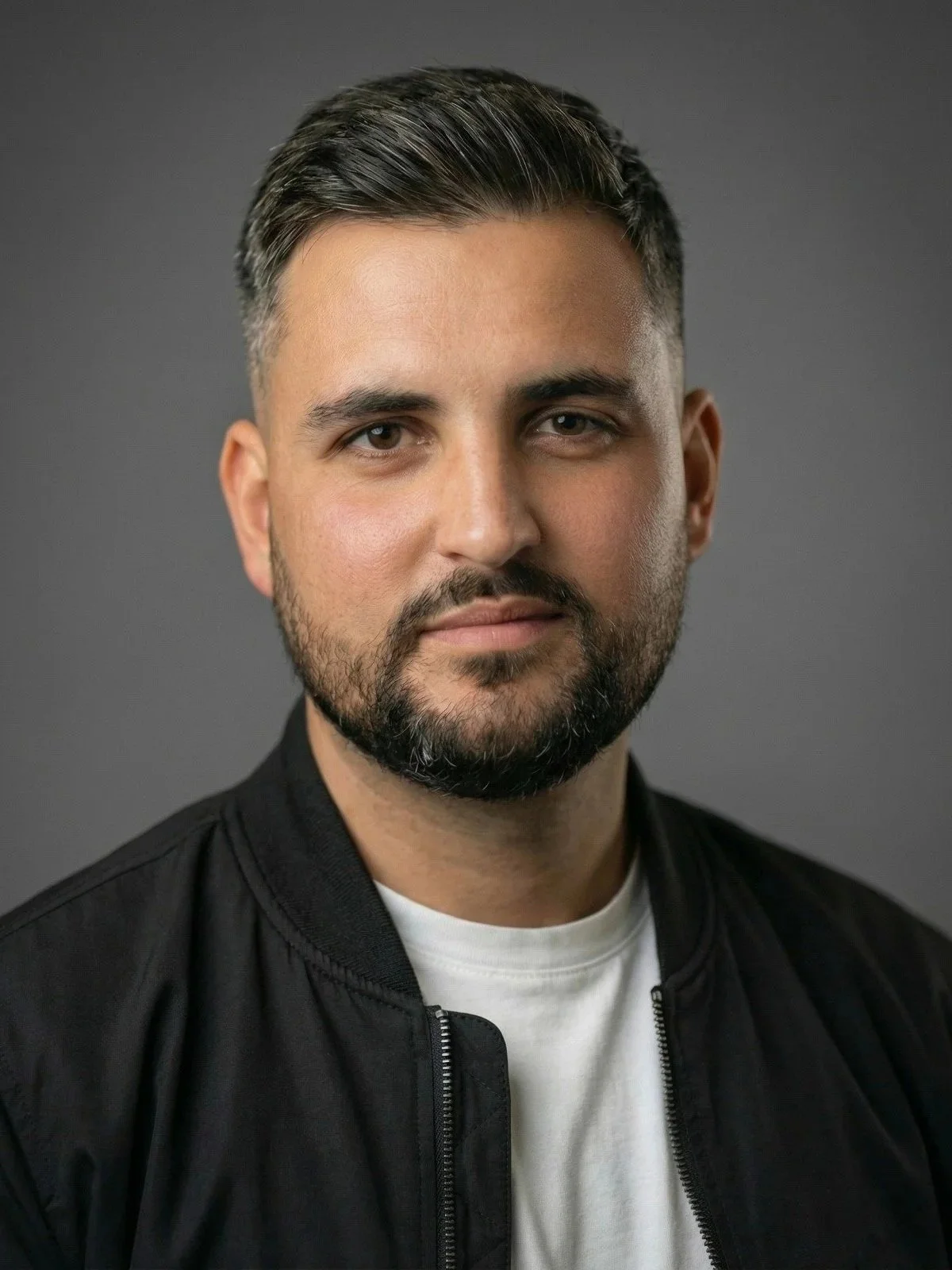Head and shoulders portrait of a man with dark hair, beard, wearing a black jacket over a white shirt against a gray background.