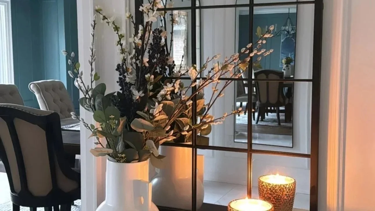 A black metal grid divider with a floral arrangement in a white vase and two candles on a dark surface, with a mirror and a dining room in the background.