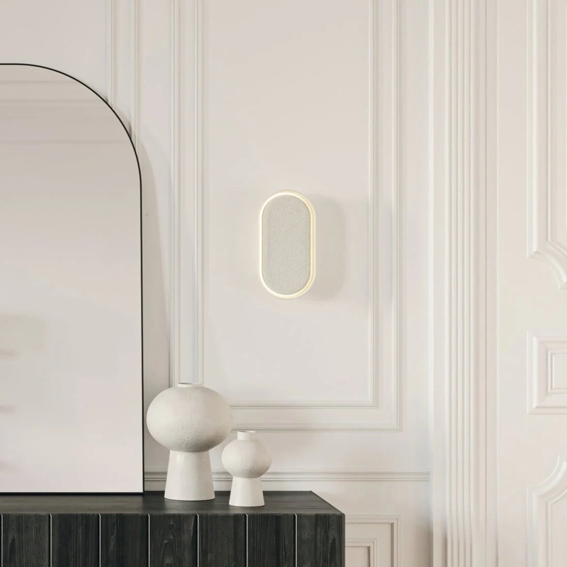 White wall with decorative molding, a wall-mounted oval speaker or light, and a dark wood sideboard with white ceramic vases.