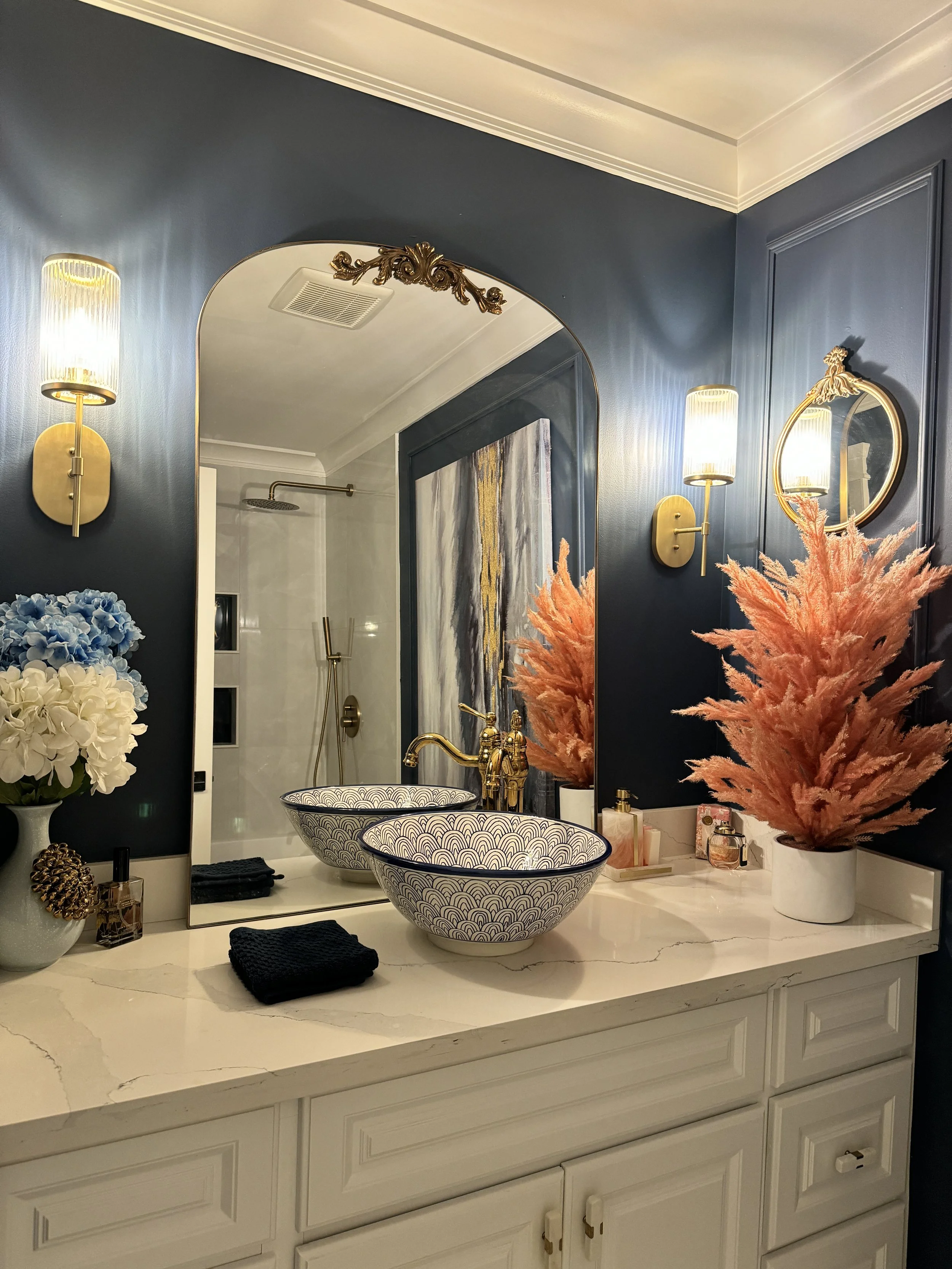Bathroom vanity with two patterned vessel sinks, a large mirror, gold fixtures, wall-mounted lights, and decorative flowers in a white vase.