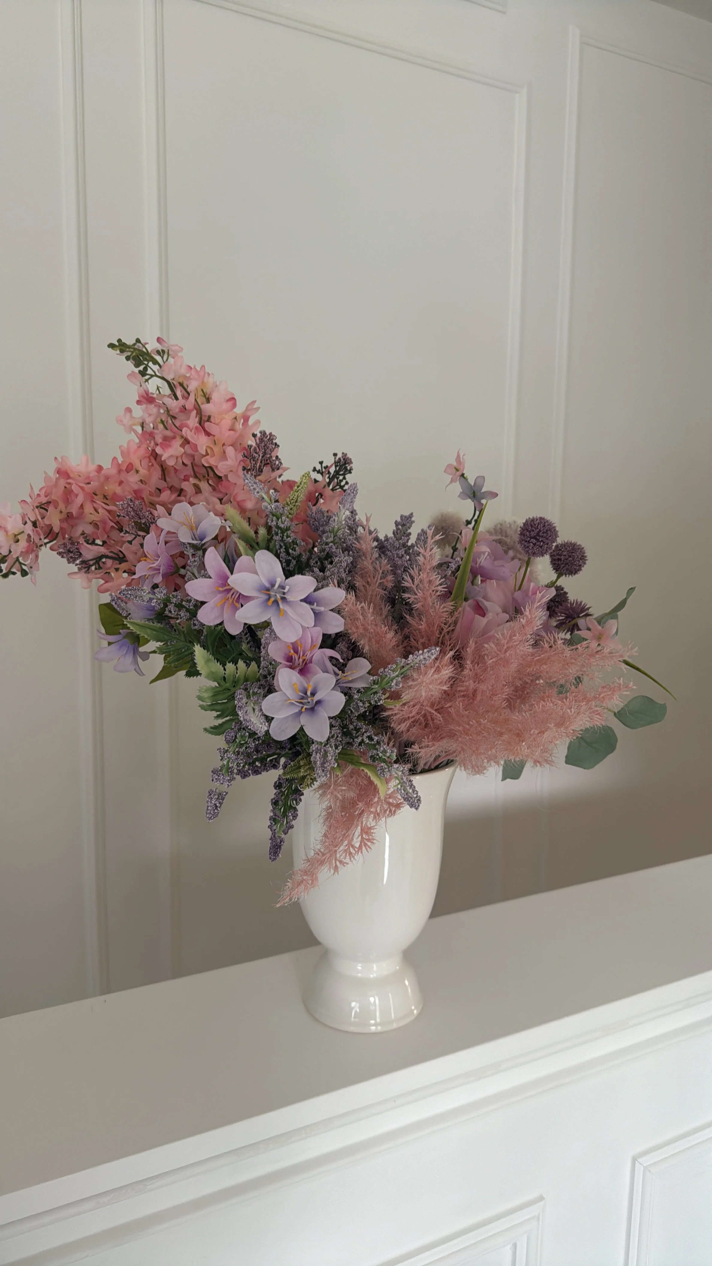 A white vase with a bouquet of pink, purple, and green flowers on a white surface against a white paneled wall.