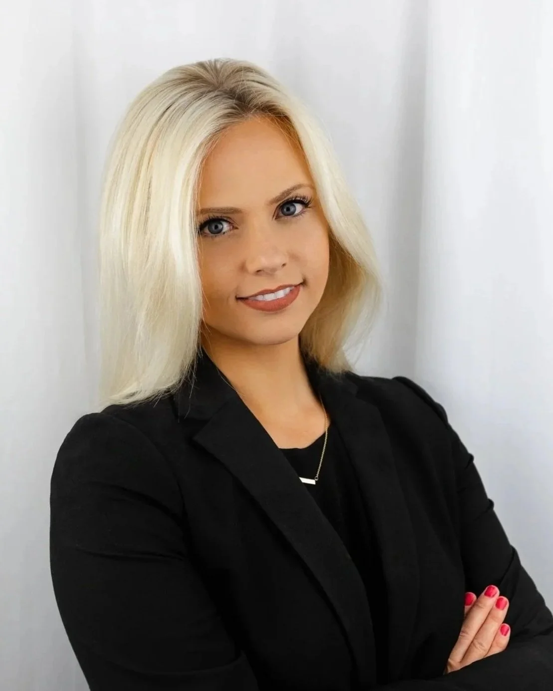 A woman with shoulder-length blonde hair and blue eyes, wearing a black blazer and a necklace, standing against a plain white background with her arms crossed.