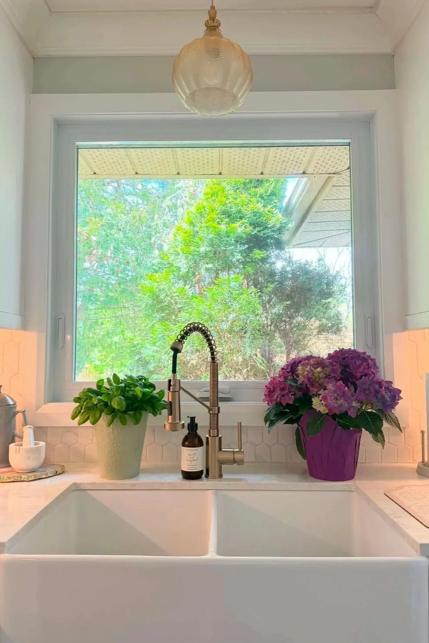 Kitchen window with two potted plants on the counter, a brown bottle soap dispenser, a kitchen faucet, and a white sink. The window reveals green trees outside. There is a purple hydrangea plant in a purple pot to the right of the sink and another green plant to the left. The background includes a white wall with honeycomb tiles.