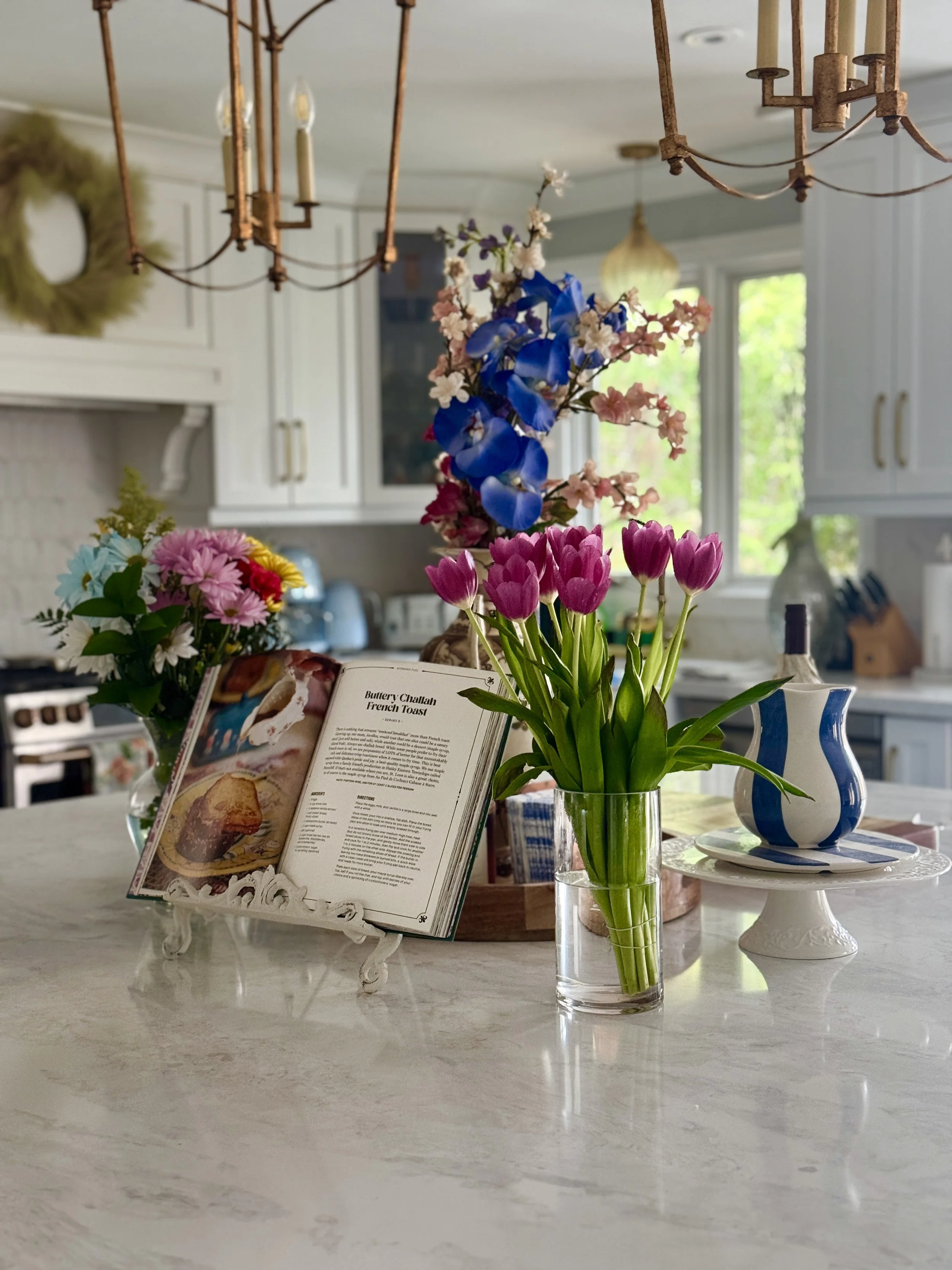 A kitchen counter with a glass vase of pink, purple, and white flowers, an open cookbook, a blue and white striped vase with pink tulips, and other decorative items in the background.