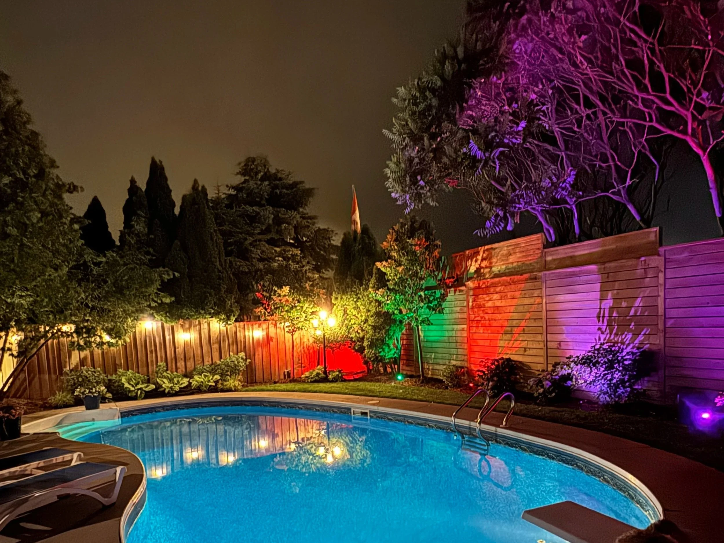 A backyard swimming pool illuminated by colorful lights, with a wooden fence, trees, and a cloudy night sky in the background.