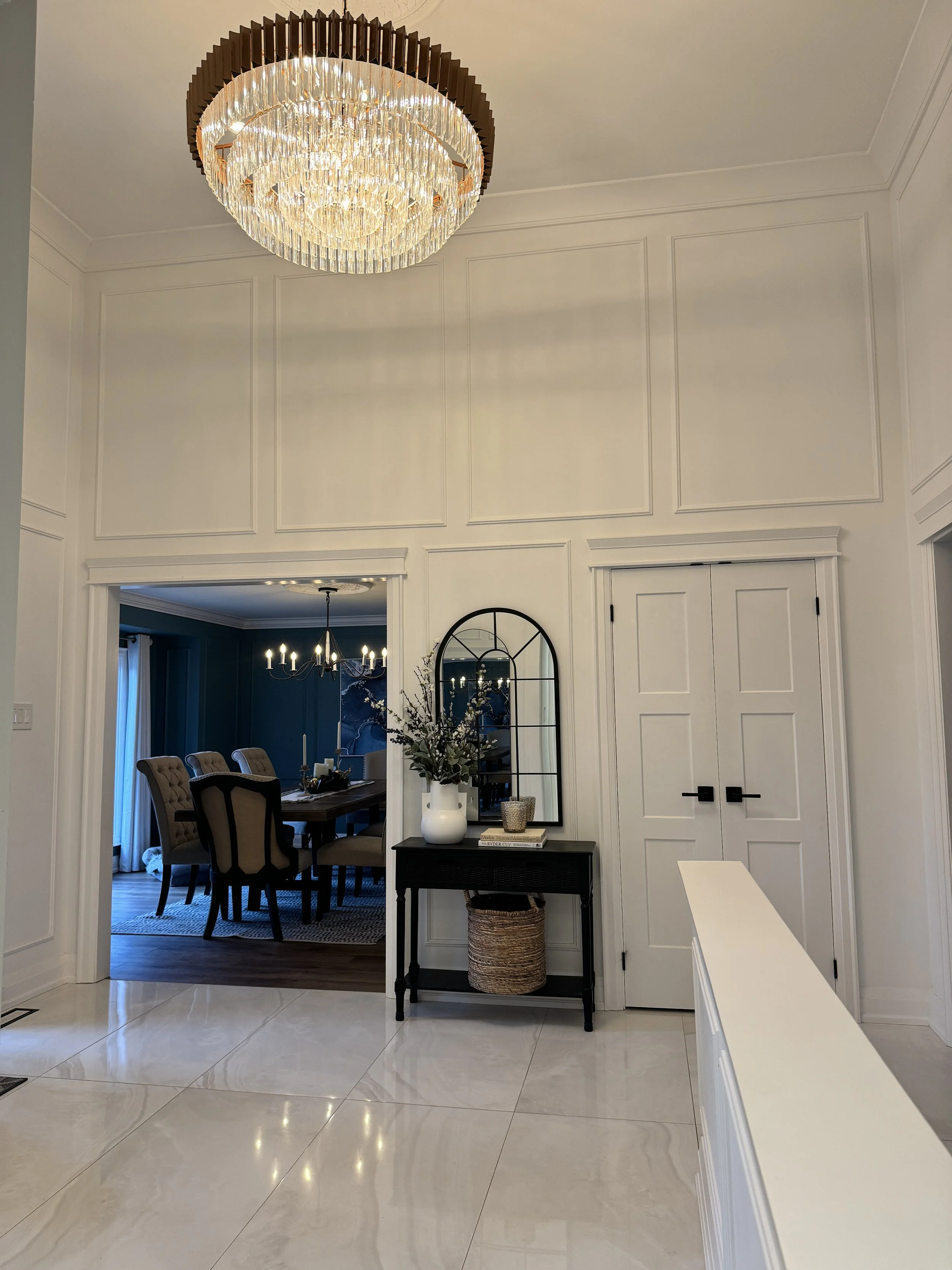 A spacious, modern dining room with a large, elegant chandelier hanging from the ceiling. The room has white walls with decorative molding, and an opening leads to a darker-colored room with a chandelier and a dining table. A small black console table holds decorative items and a plant, with a woven basket underneath. The floor is tiled with shiny, large marble-like tiles.