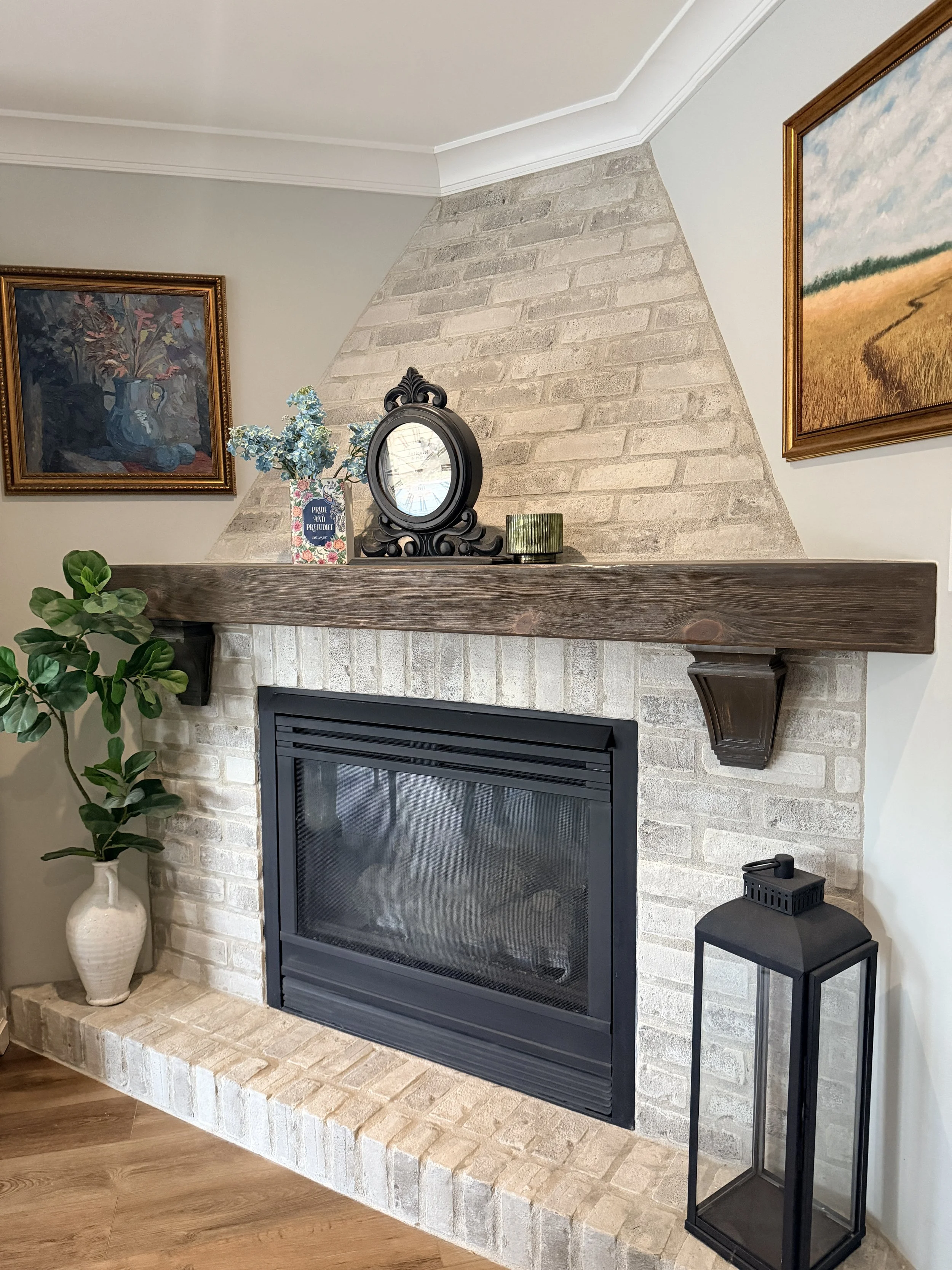 Fireplace with a brick surround and a wooden mantel decorated with a vase of blue flowers, a mirror, and a green candle. Two framed paintings hang above, a large houseplant in a white vase is on the left, and a black lantern-style decorative piece sits on a small table on the right.