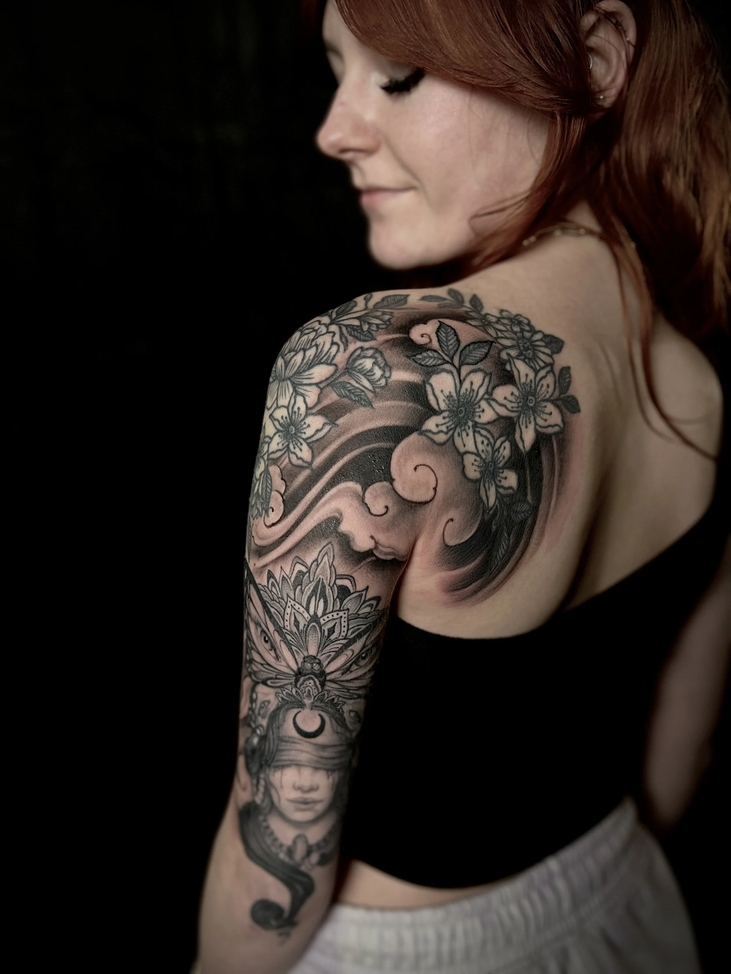 A woman with red hair and tattoos on her shoulder and arm, including flowers, a face with a crescent moon on the forehead, and swirling patterns, against a black background.