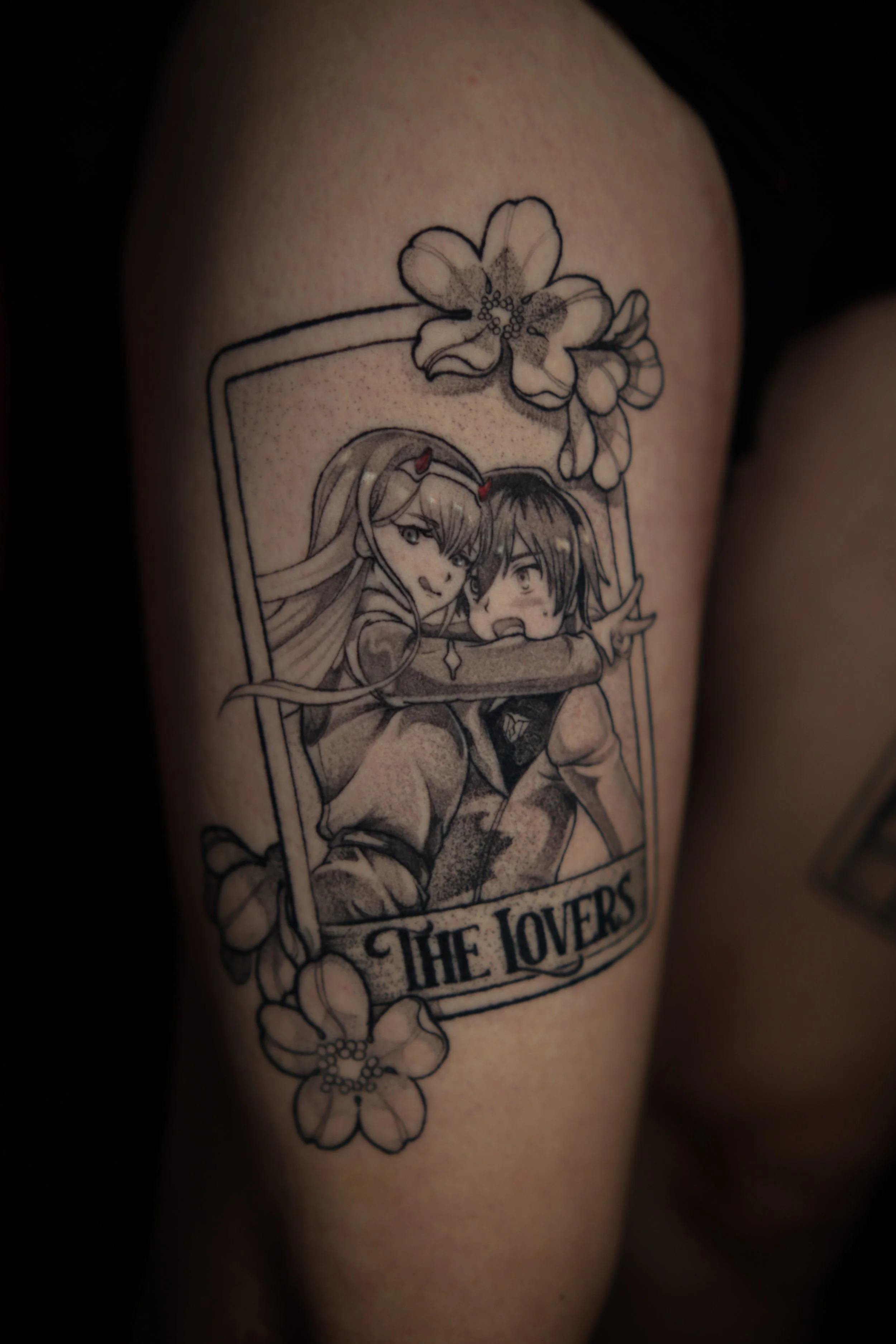 Black and gray tattoo on person's arm depicting a male and female anime-style characters inside a frame, with flowers surrounding the frame, and the text "THE LOVERS" at the bottom.