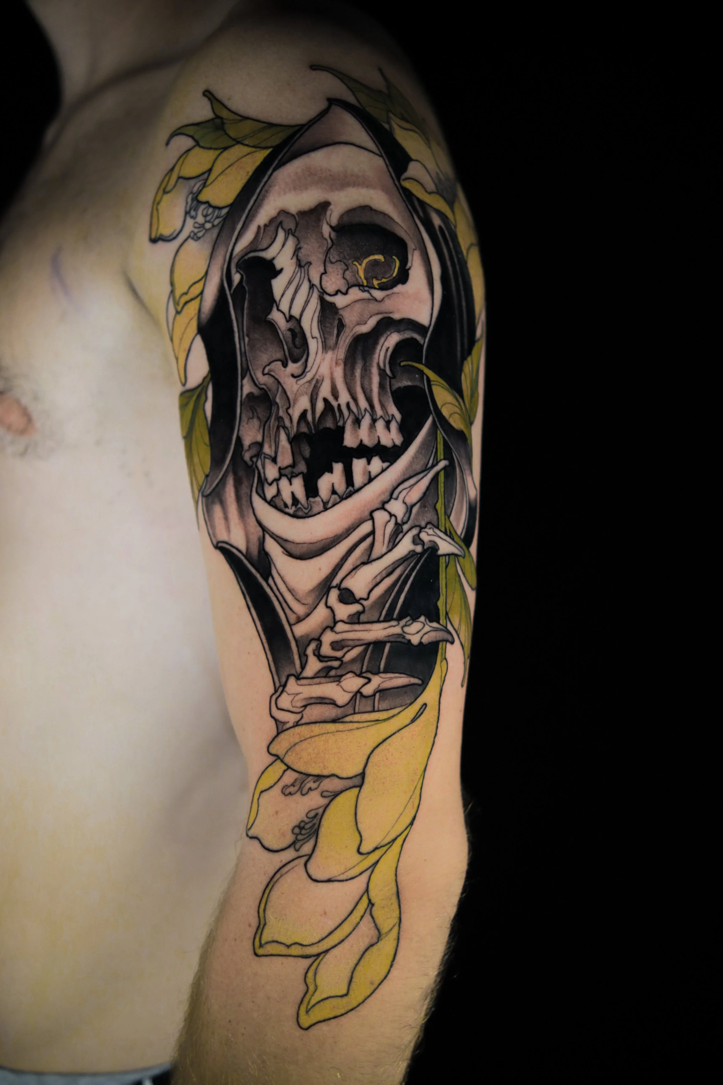 Tattoo of a skull with a finger pointing, surrounded by yellow leaves on an upper arm.
