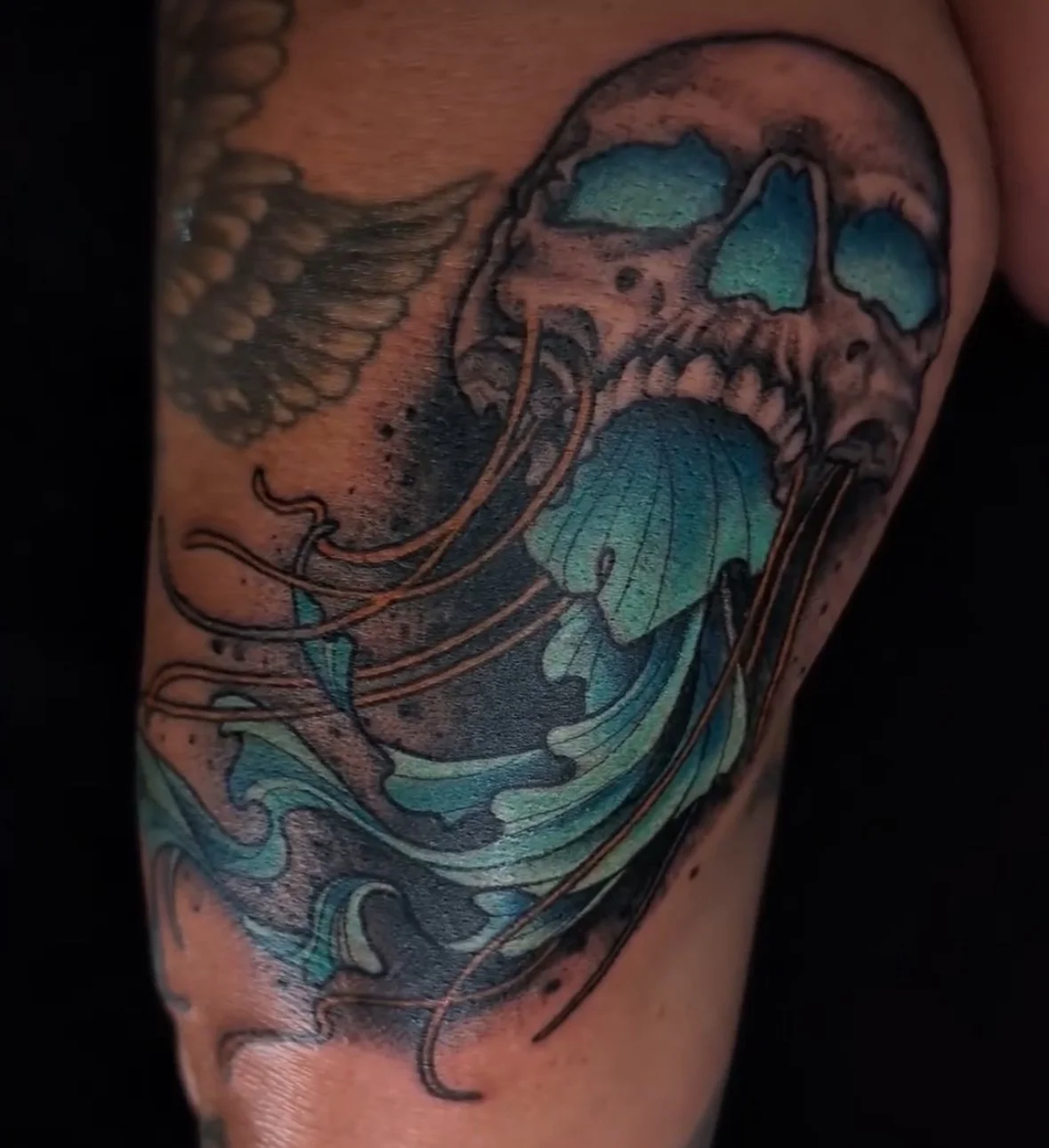 Tattoo of a skull with flowing hair and waves, rendered in blue and green hues.