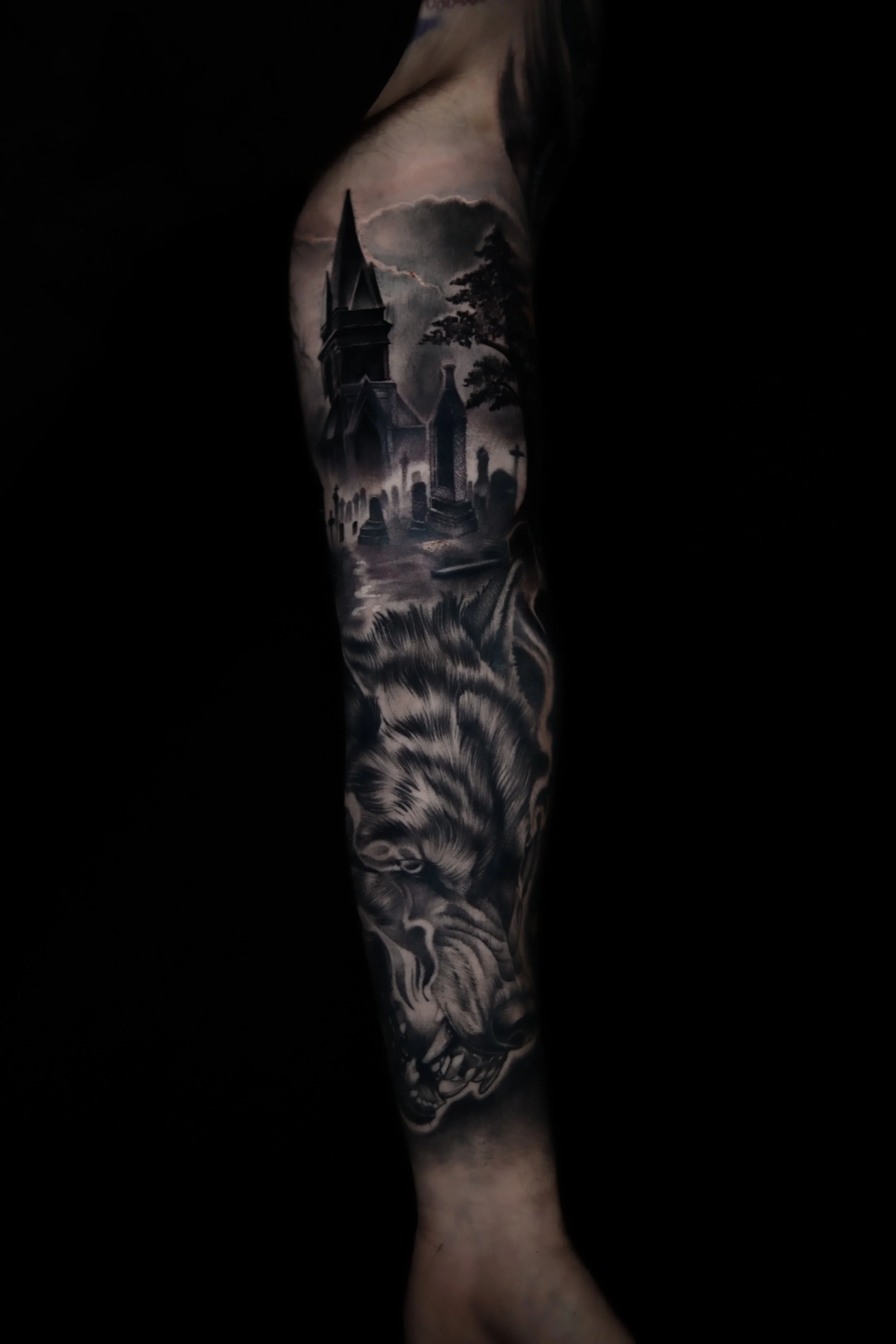 A black and grey tattoo sleeve depicting a haunted house with a graveyard in the background and a fierce-looking wolf in the foreground.