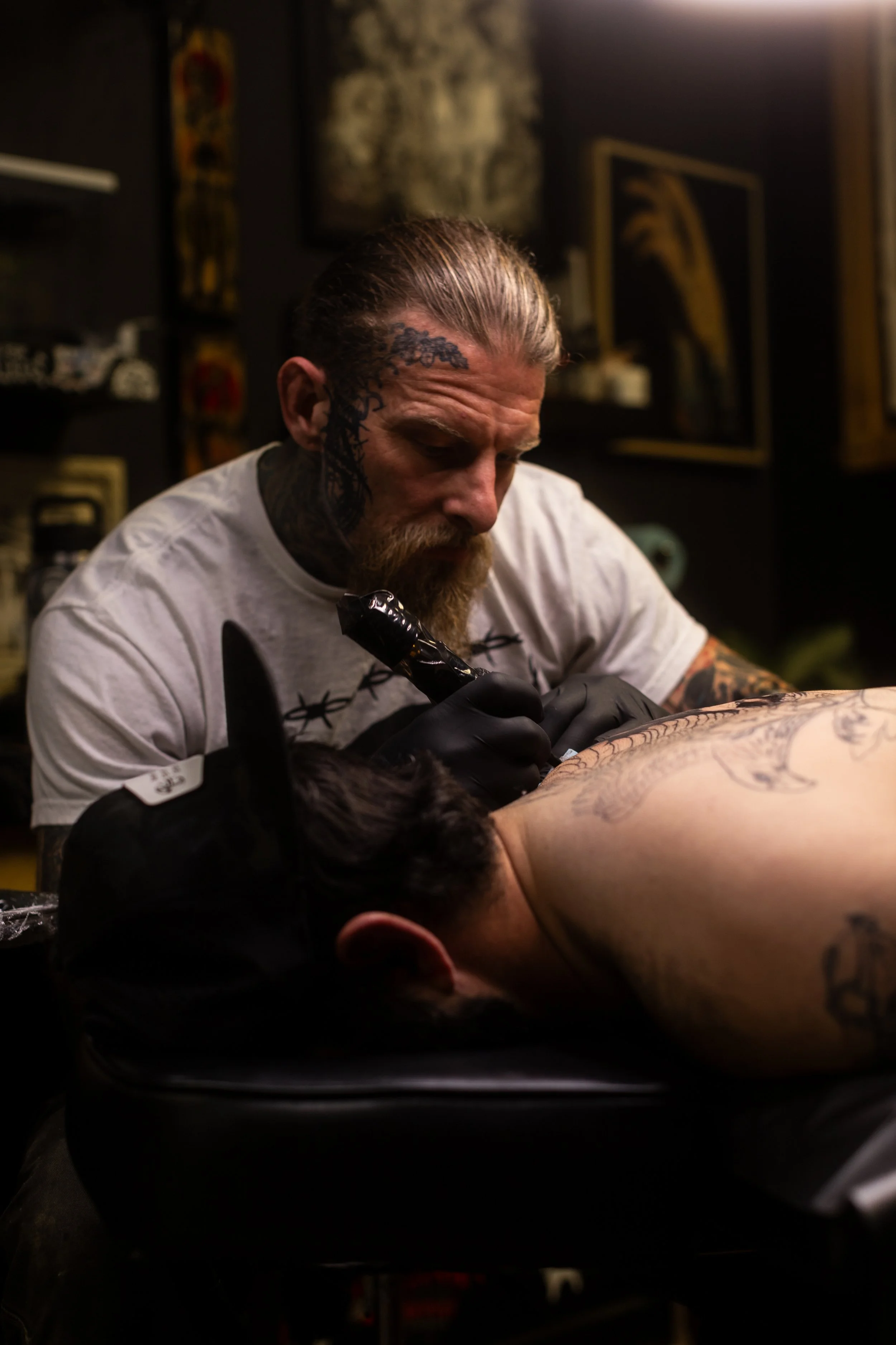 Tattoo artist with tattoos and a beard creating a tattoo on a person's back.