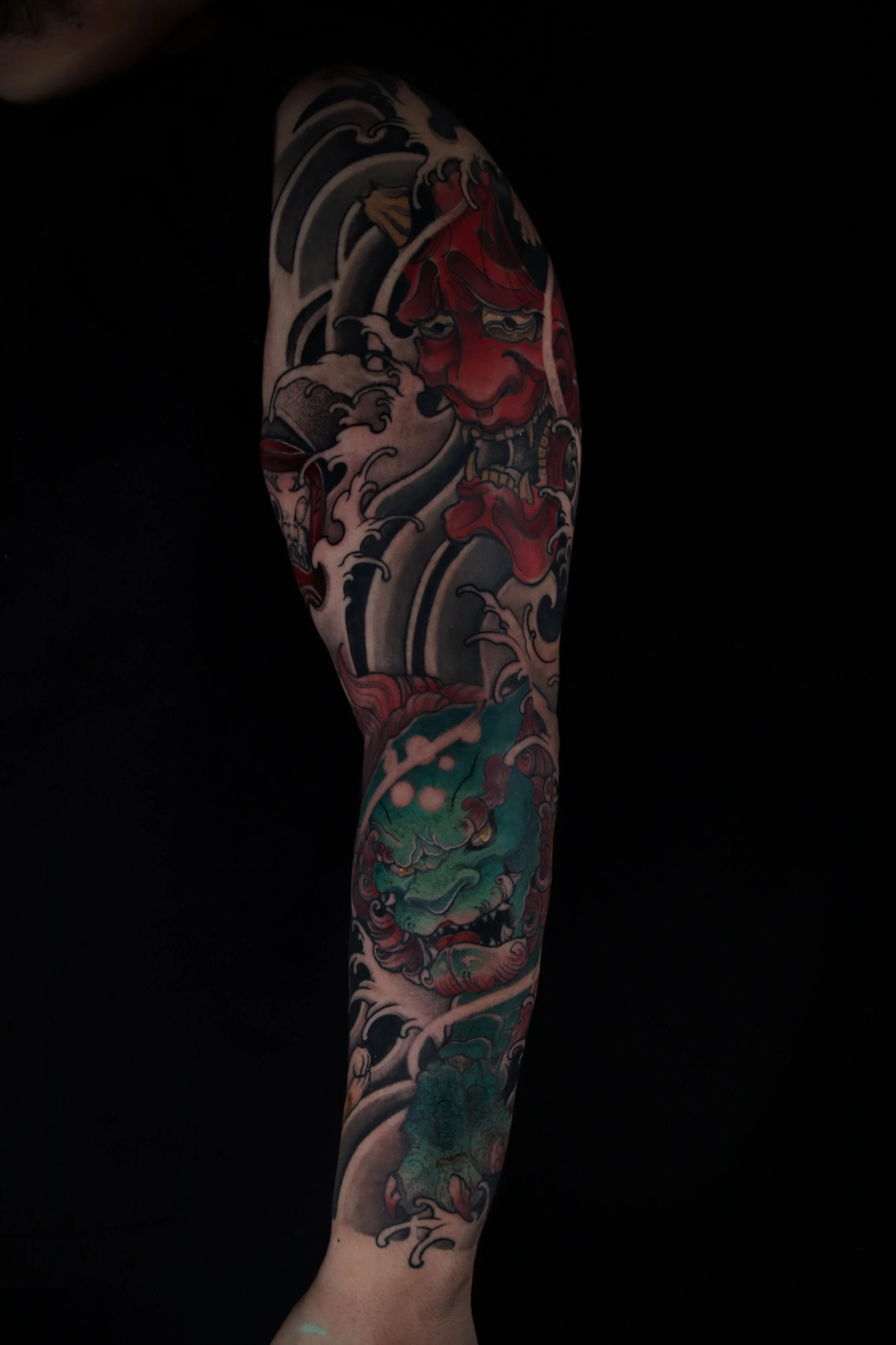Detailed Japanese-style tattoo sleeve featuring a red oni mask, waves, and a green creature on the arm.