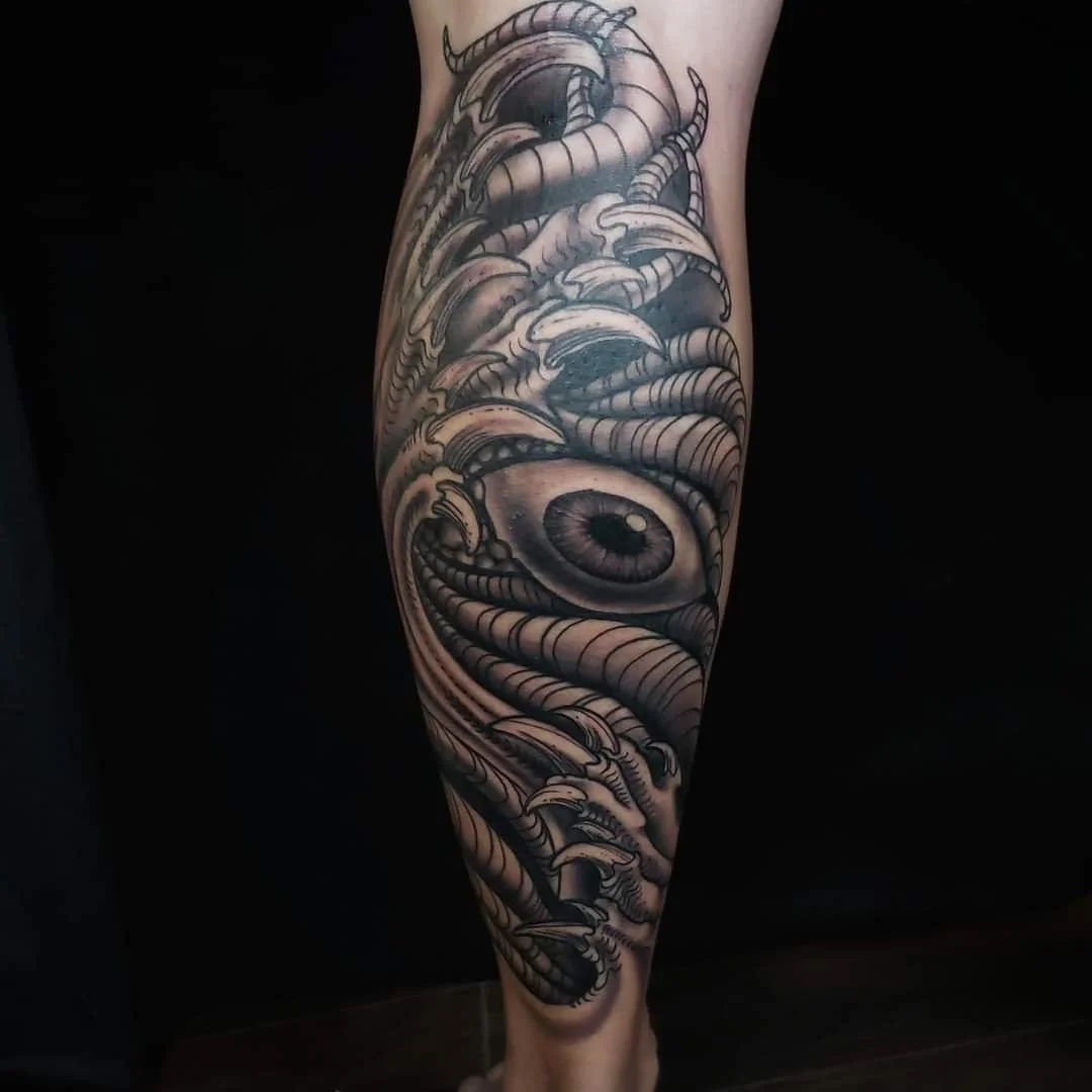 Detailed black and gray tattoo of a giant eye, surrounded by swirling abstract shapes and patterns on a person's leg.