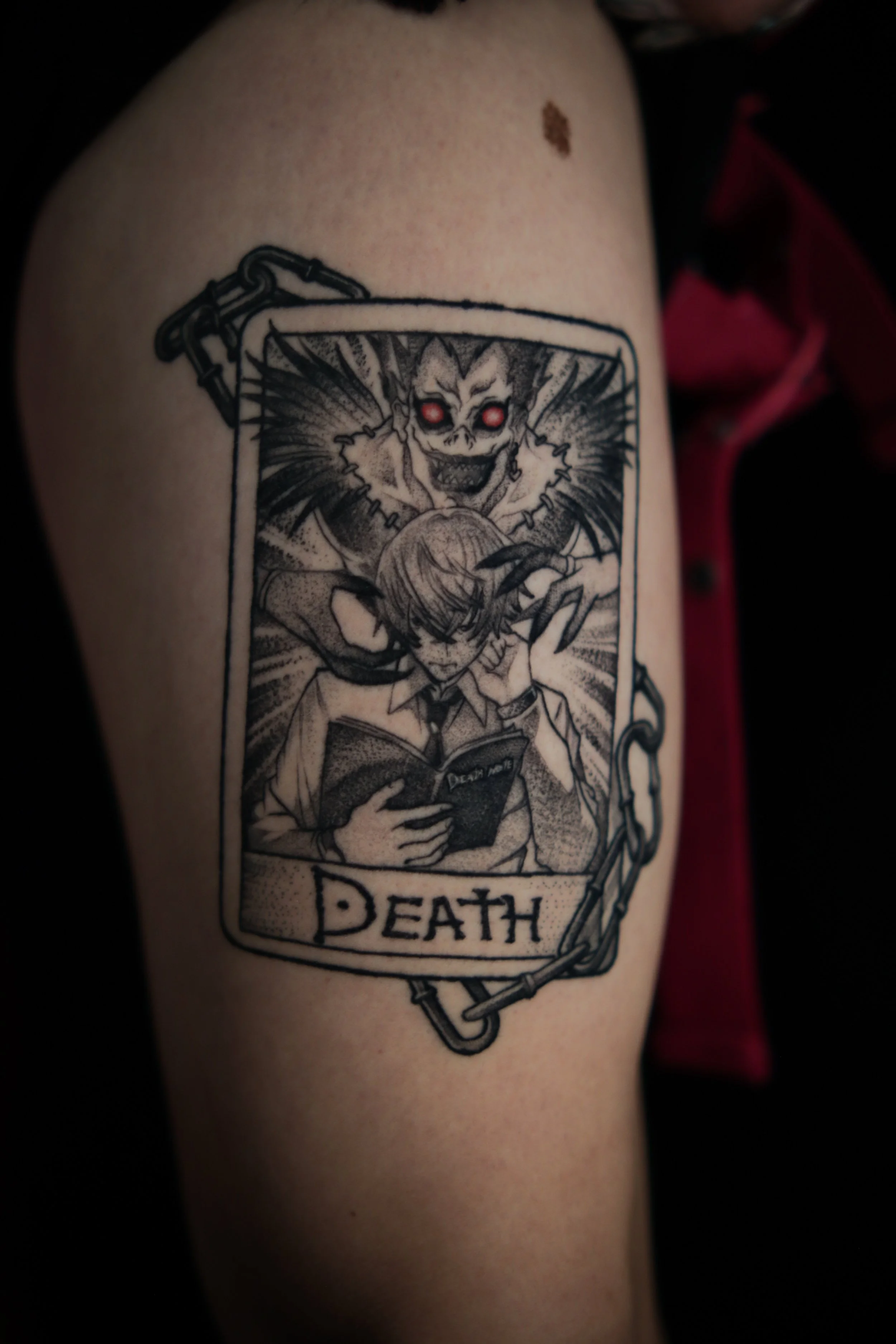 Tattoo of a tarot card titled 'DEATH,' featuring a sinister skull with glowing red eyes above a person reading a book, with a demonic creature with sharp claws in the background.