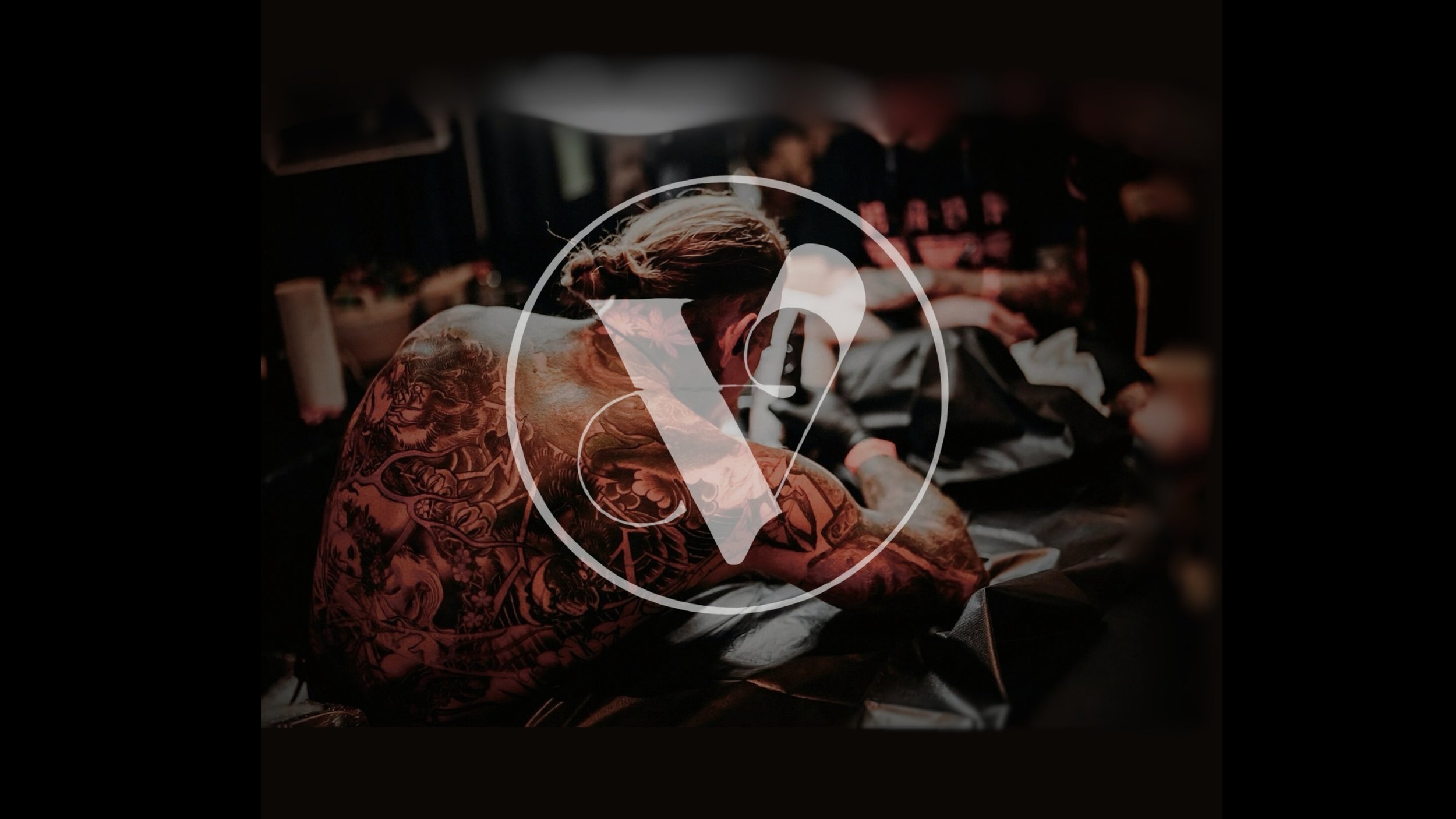 A person with tattoos on their back and arm, sitting at a barbershop chair, getting a haircut from a barber. Others are visible in the background. The image is overlaid with a transparent logo featuring a V inside a circle.