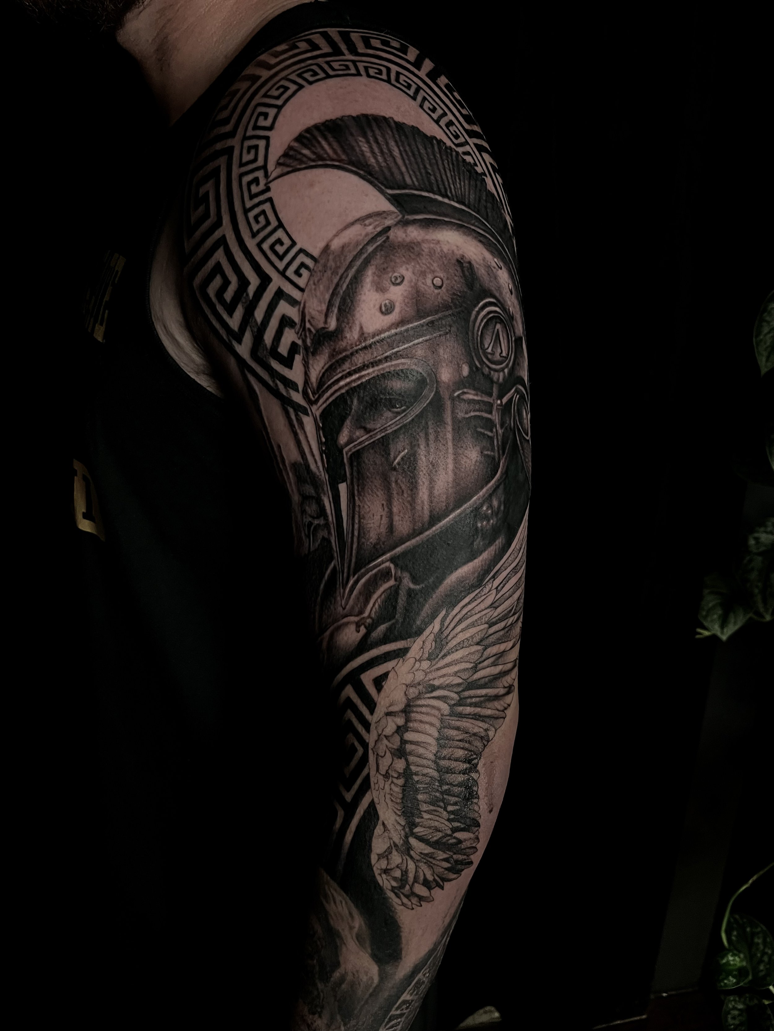 Tattoo of a Spartan helmet with detailed shading, surrounded by a circular pattern and a winged creature on the arm.