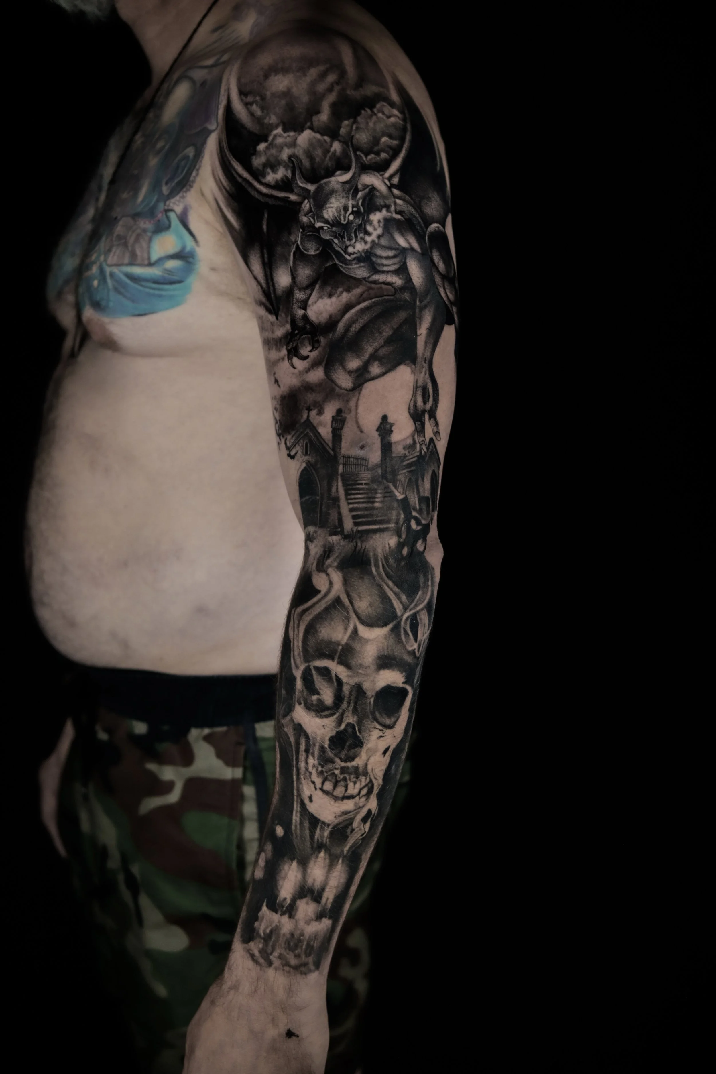 Tattooed arm with designs of a skull, a demon, clouds, and buildings, against a black background.