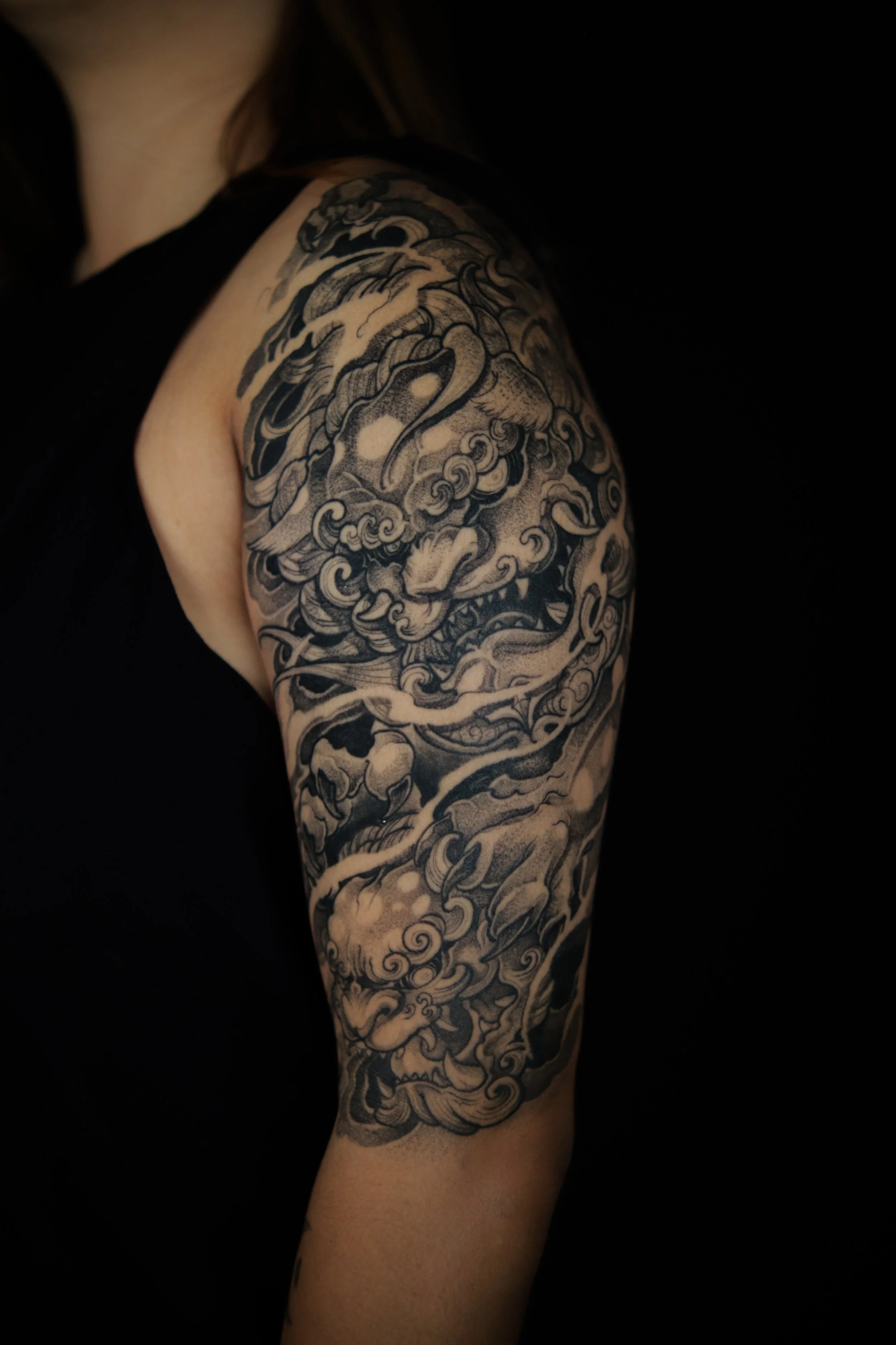 Black and gray tattoo of a fierce Japanese-style lion or mythical creature on a person's upper arm and shoulder, surrounded by swirling clouds and flames.