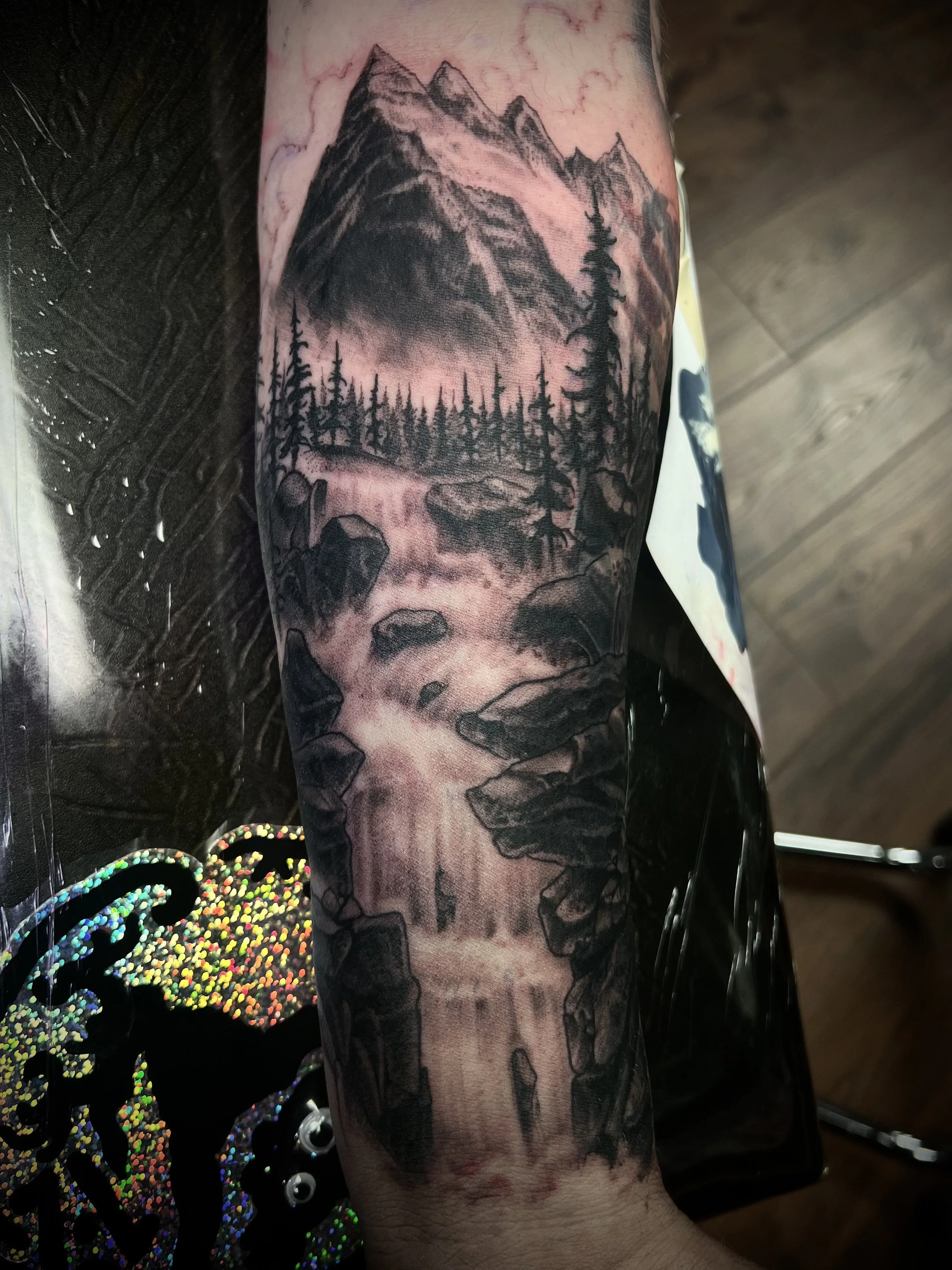 Tattoo of a mountain landscape with forest, a river, and a skull at the bottom, rendered in black ink.