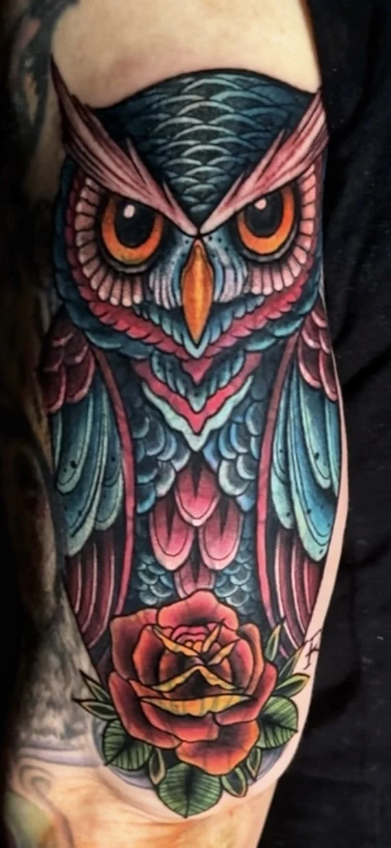 Colorful owl tattoo on person's upper arm with a rose at the bottom.