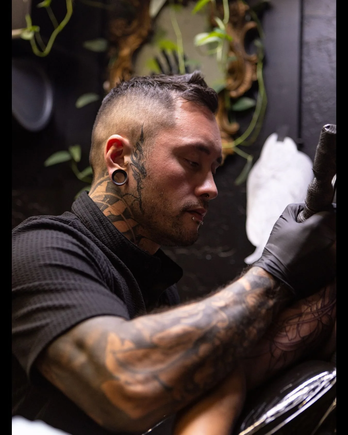 Tattooed man with gauged ears and facial piercings, tattooed arm, and wearing black gloves, working on a tattoo in a studio with tattoo equipment and plant decorations in the background.