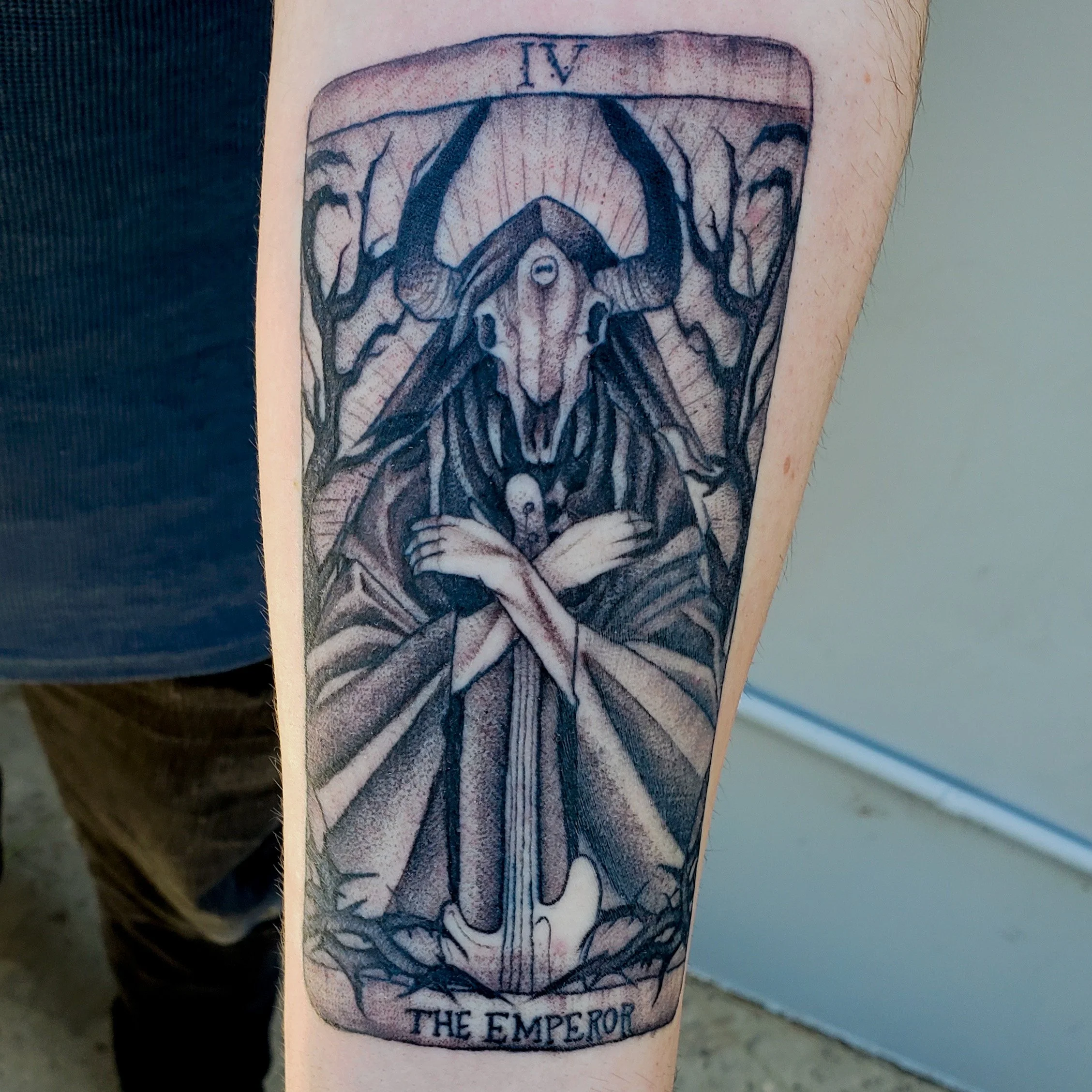 Tattoo of a tarot card depicting The Emperor, featuring a ram skull with horns, a figure with a beard, crossed arms, and a crown, surrounded by trees, with the text 'THE EMPEROR' at the bottom.
