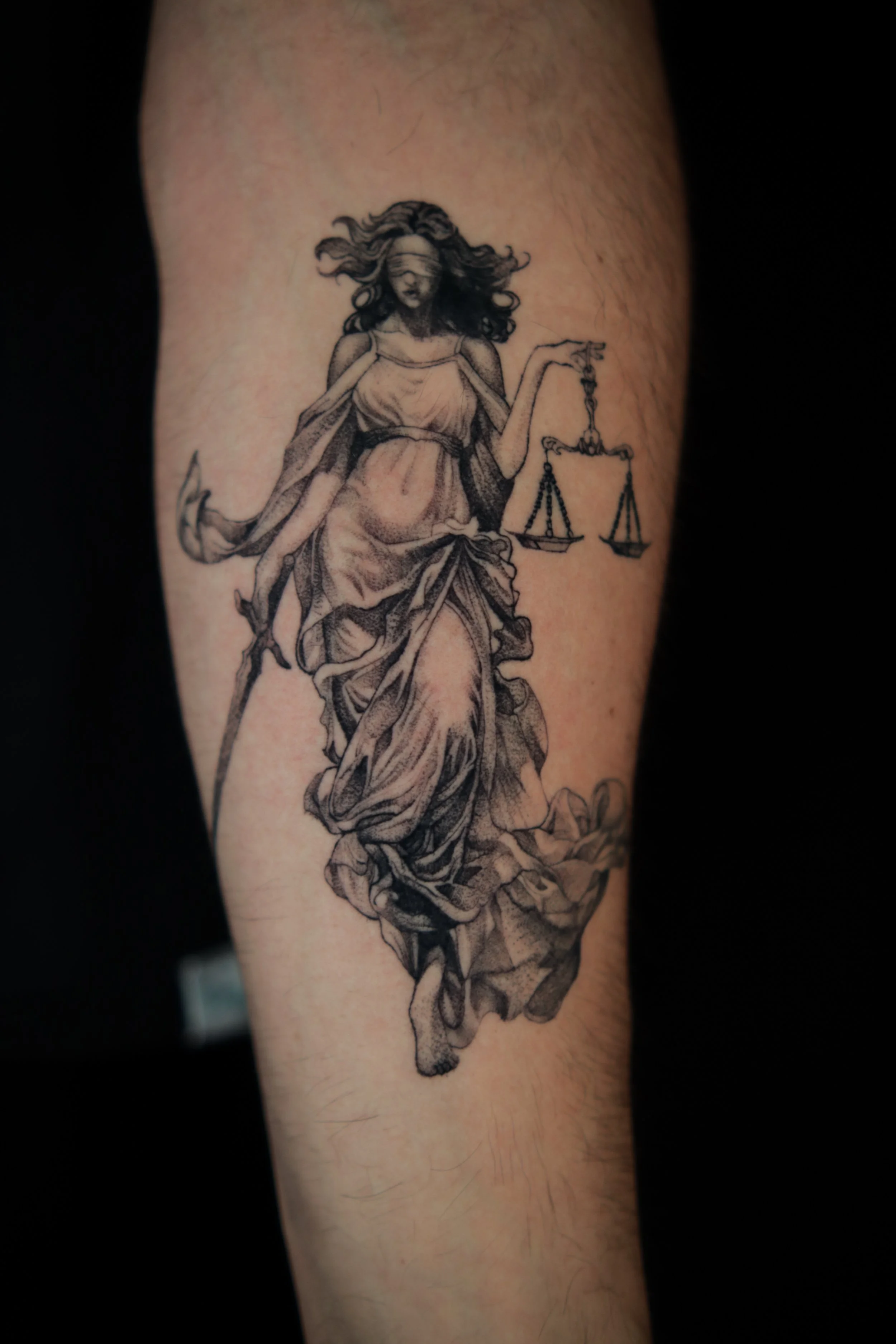 Black and grey tattoo of Lady Justice with a blindfold, holding a scale in her left hand and a sword in her right hand, wearing flowing robes.