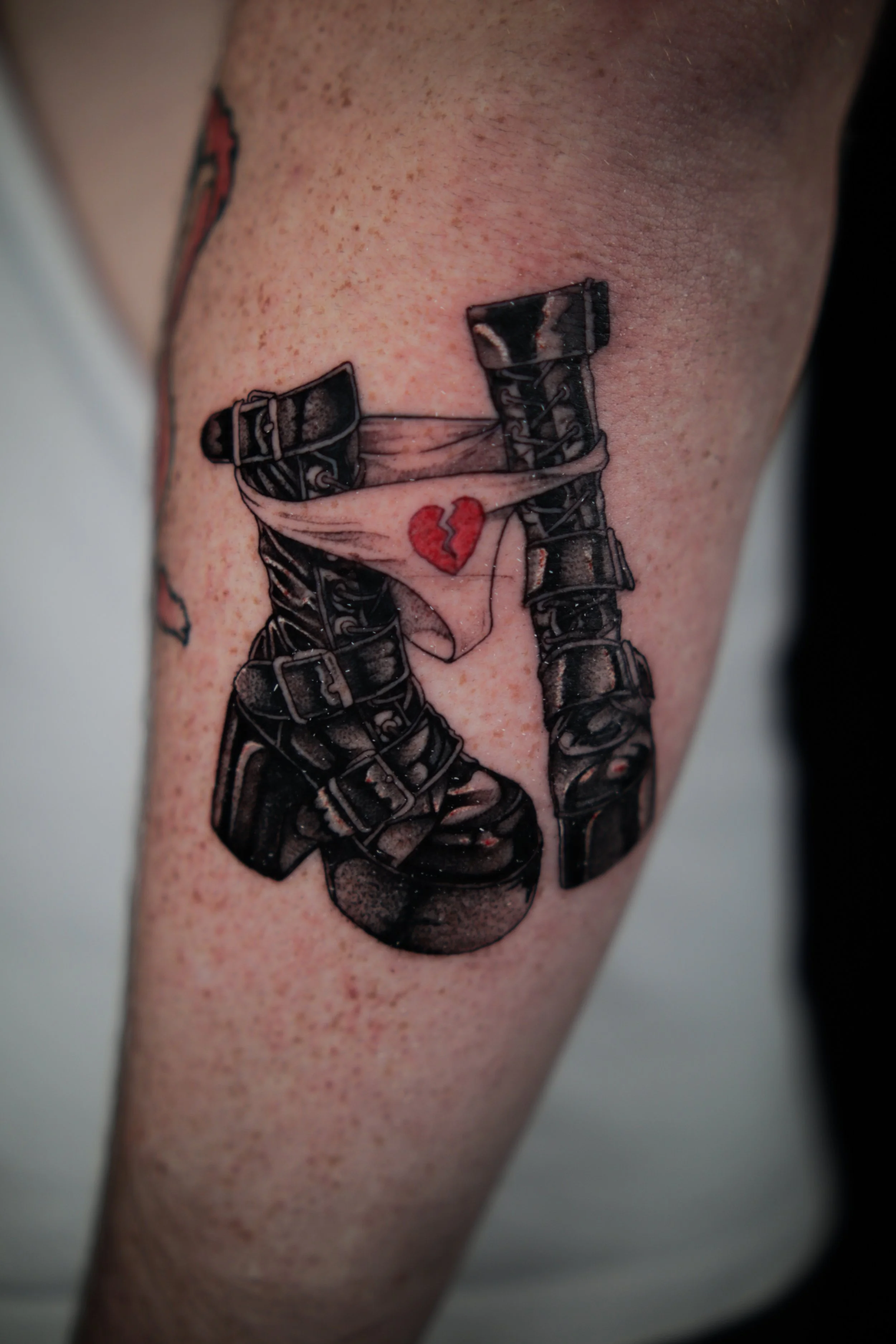 Tattoo of a pair of black combat boots with a red broken heart in between, on the arm.