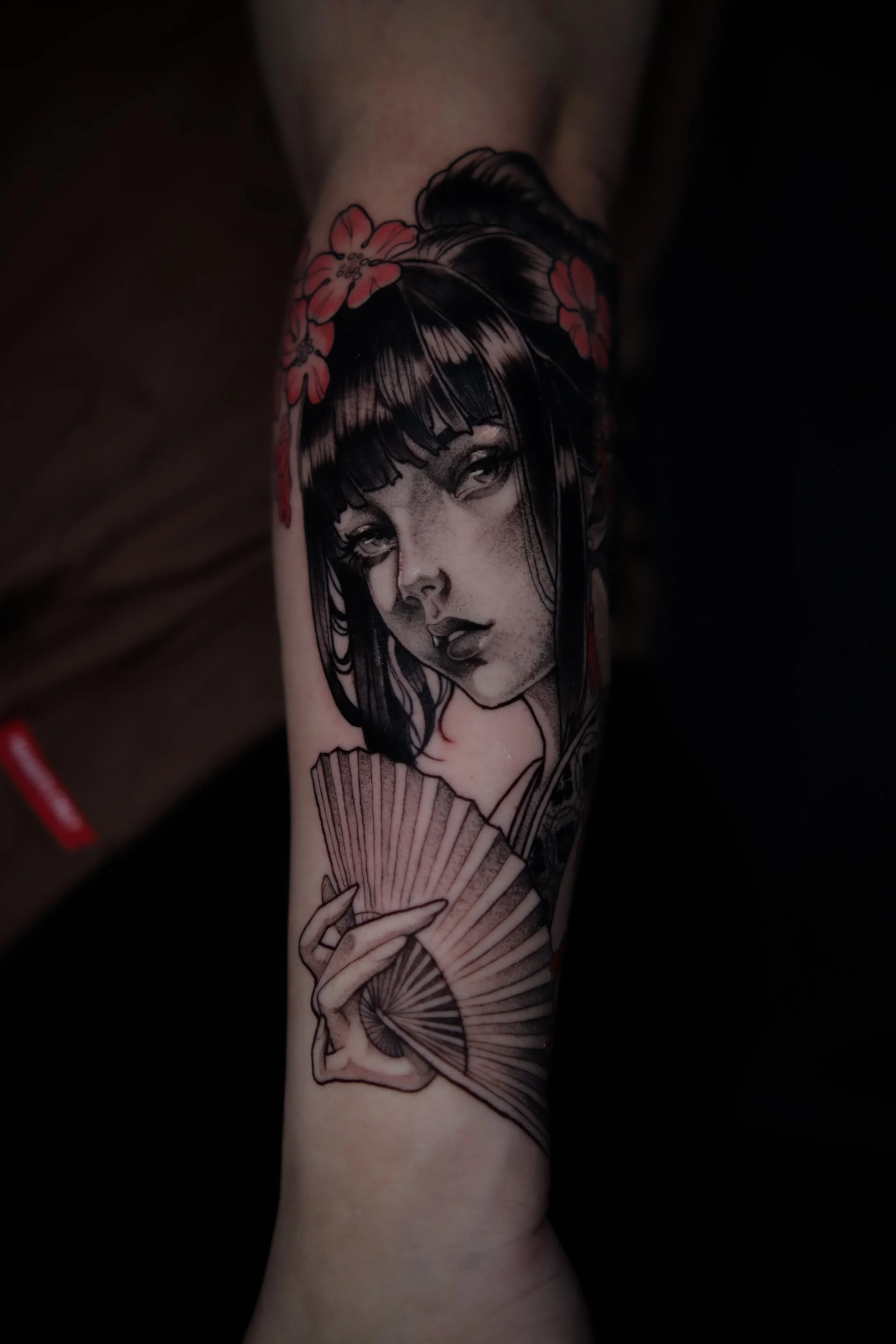 Tattoo of a woman with dark hair and bangs holding a pink folding fan, adorned with pink flowers in her hair.