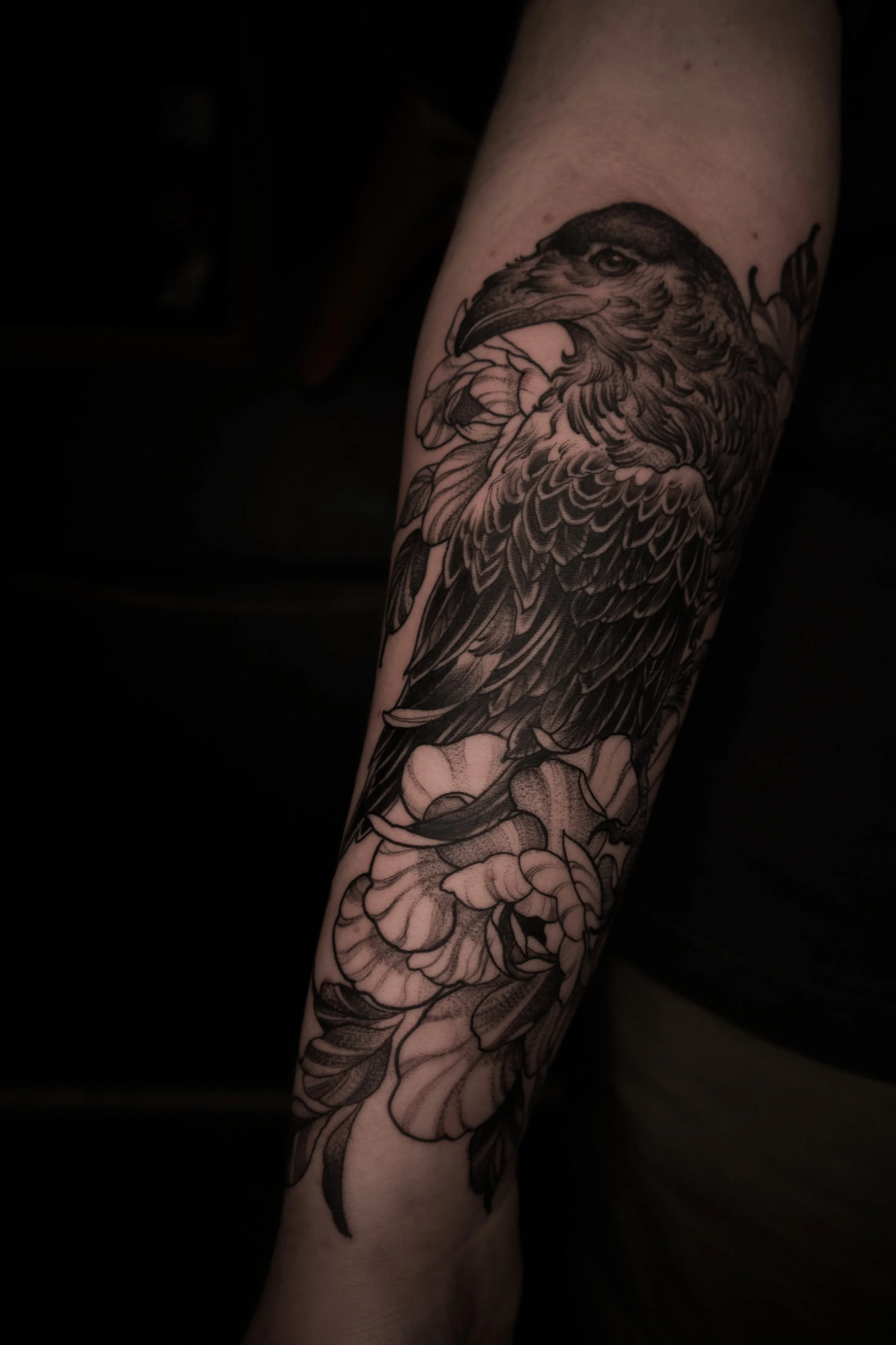Black and gray tattoo of a bird, likely an eagle, with detailed feathers, perched among flowers on a person's forearm.