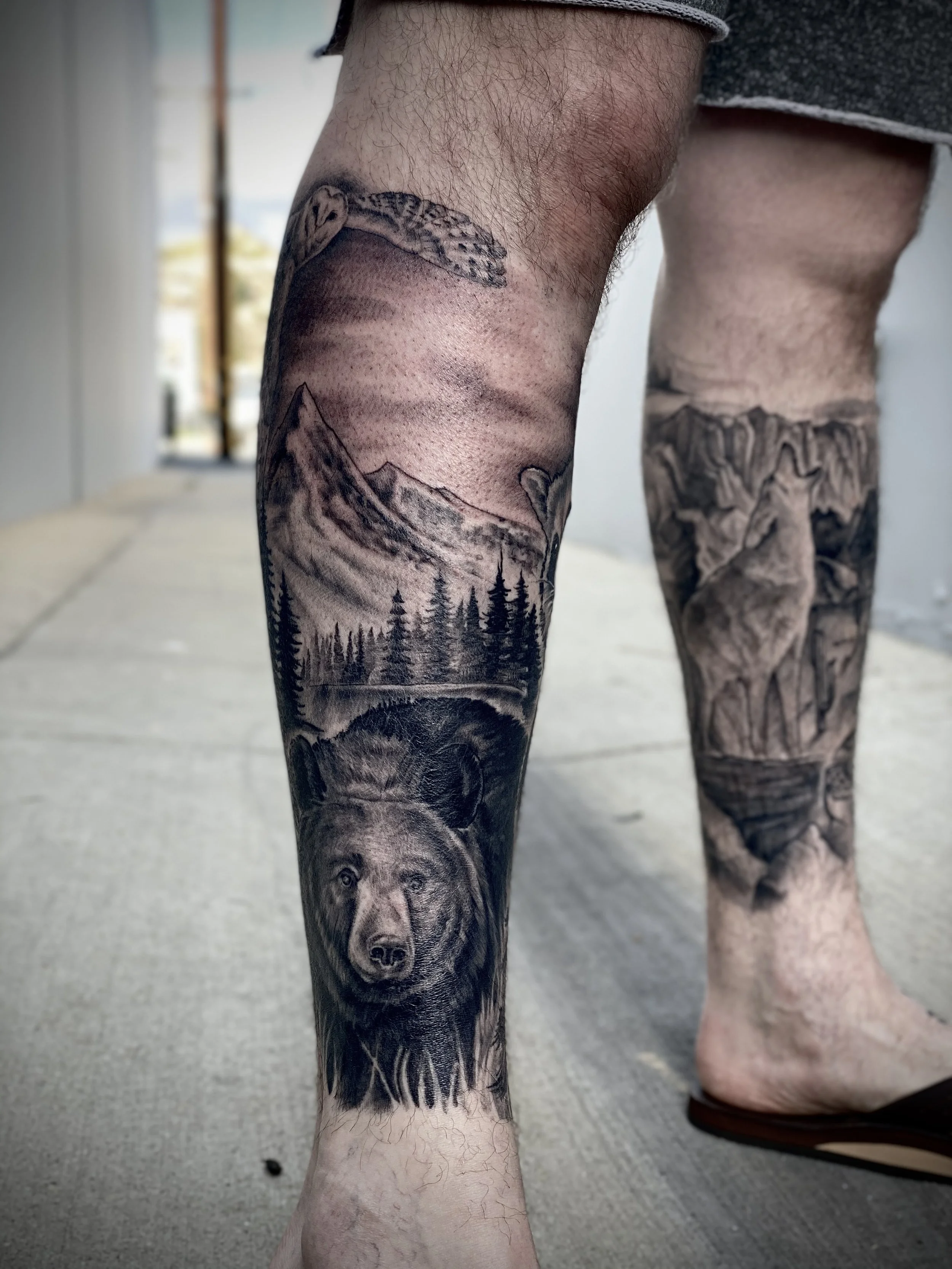 Tattoo of a bear's face on a person's lower leg, with a mountain lake scene in the background.