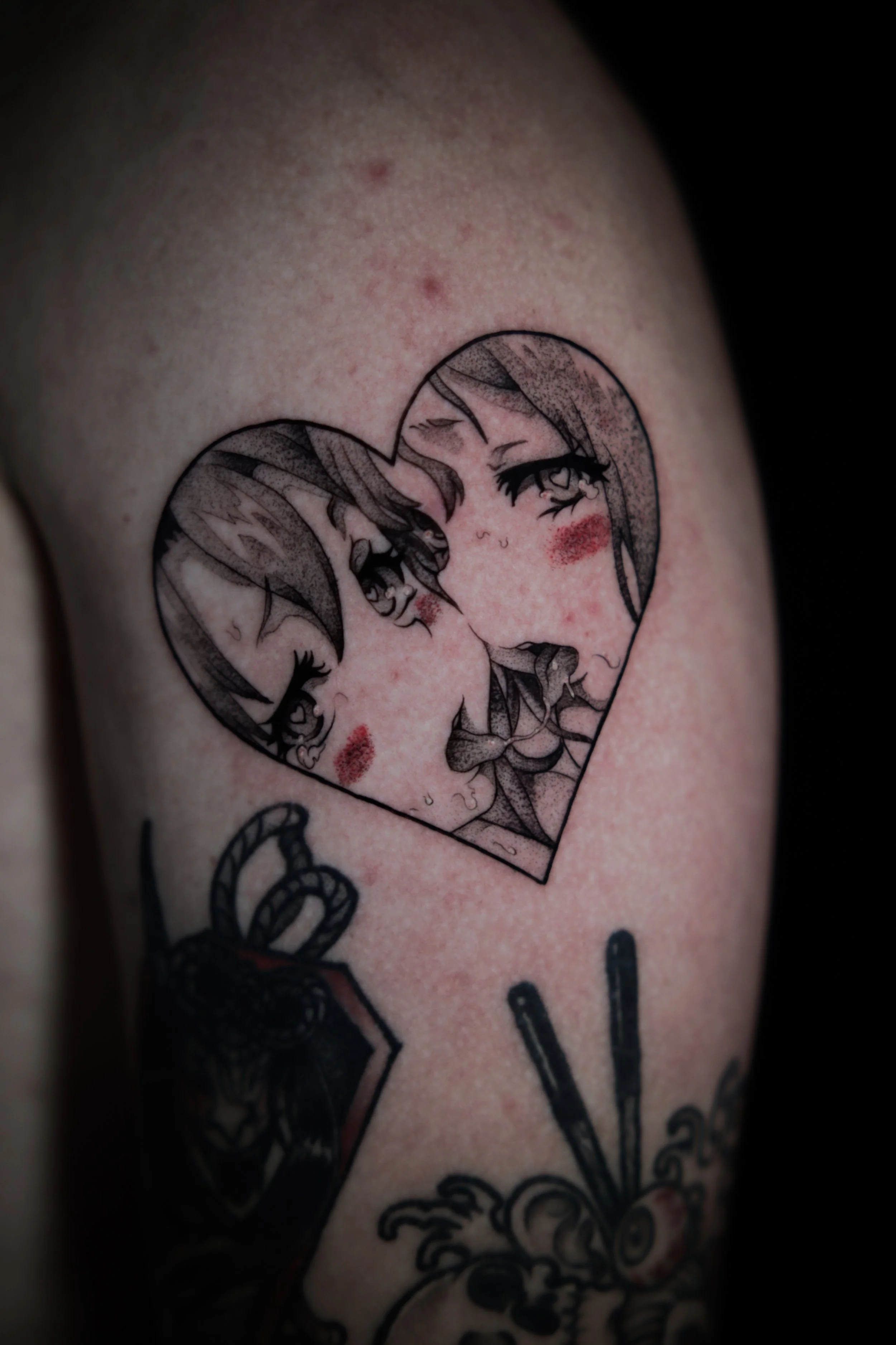 Tattoo of two anime-style female faces inside a heart outline with blush on their cheeks.