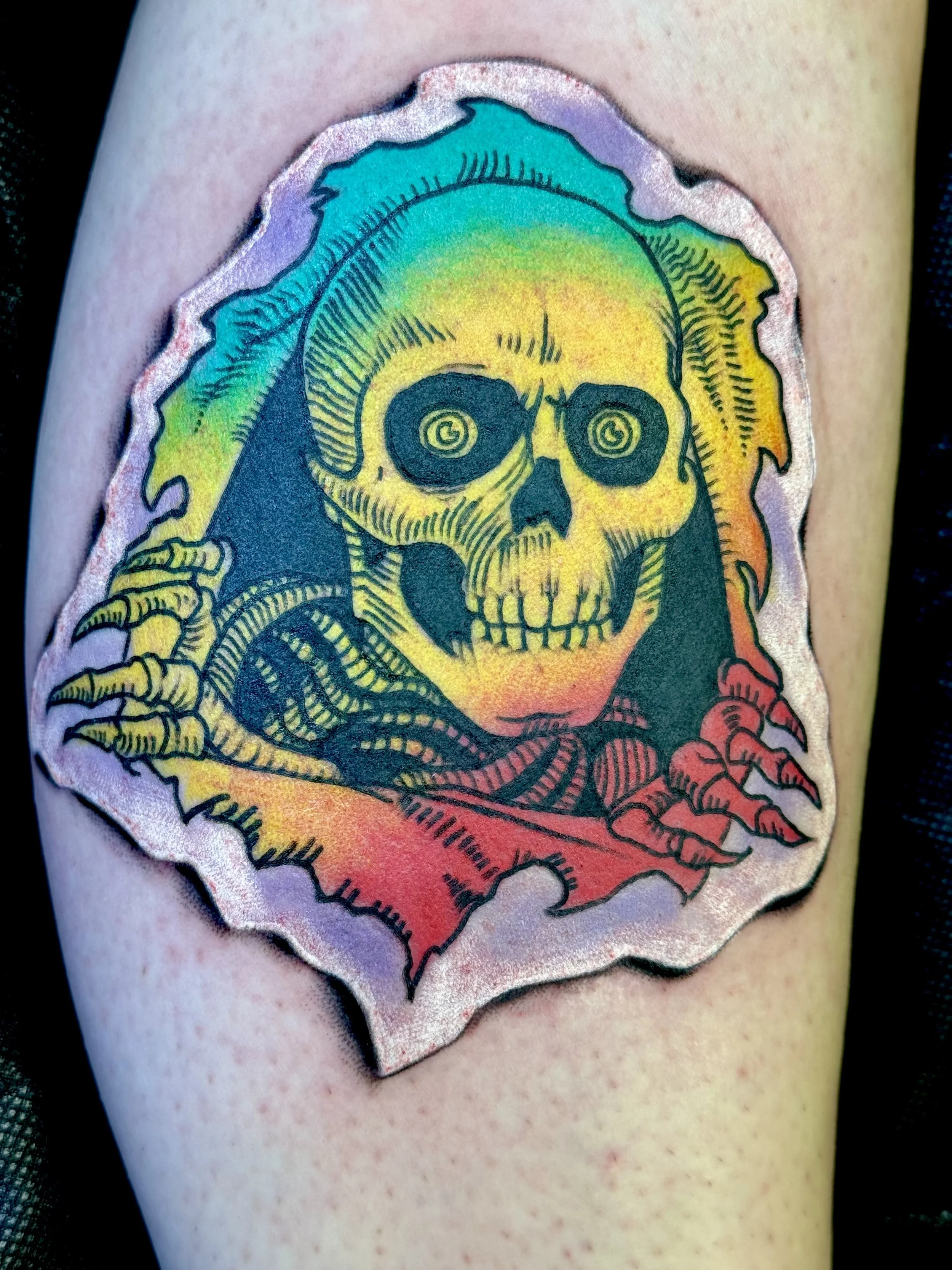 Colorful tattoo of a skull with spiral eyes, surrounded by rainbow-colored flames, with skeletal hands reaching out.