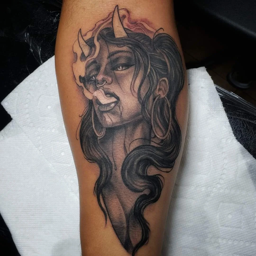 Black and gray tattoo of a woman with long flowing hair and unicorn features, including a horn on her forehead, hoop earrings, and a fierce expression.
