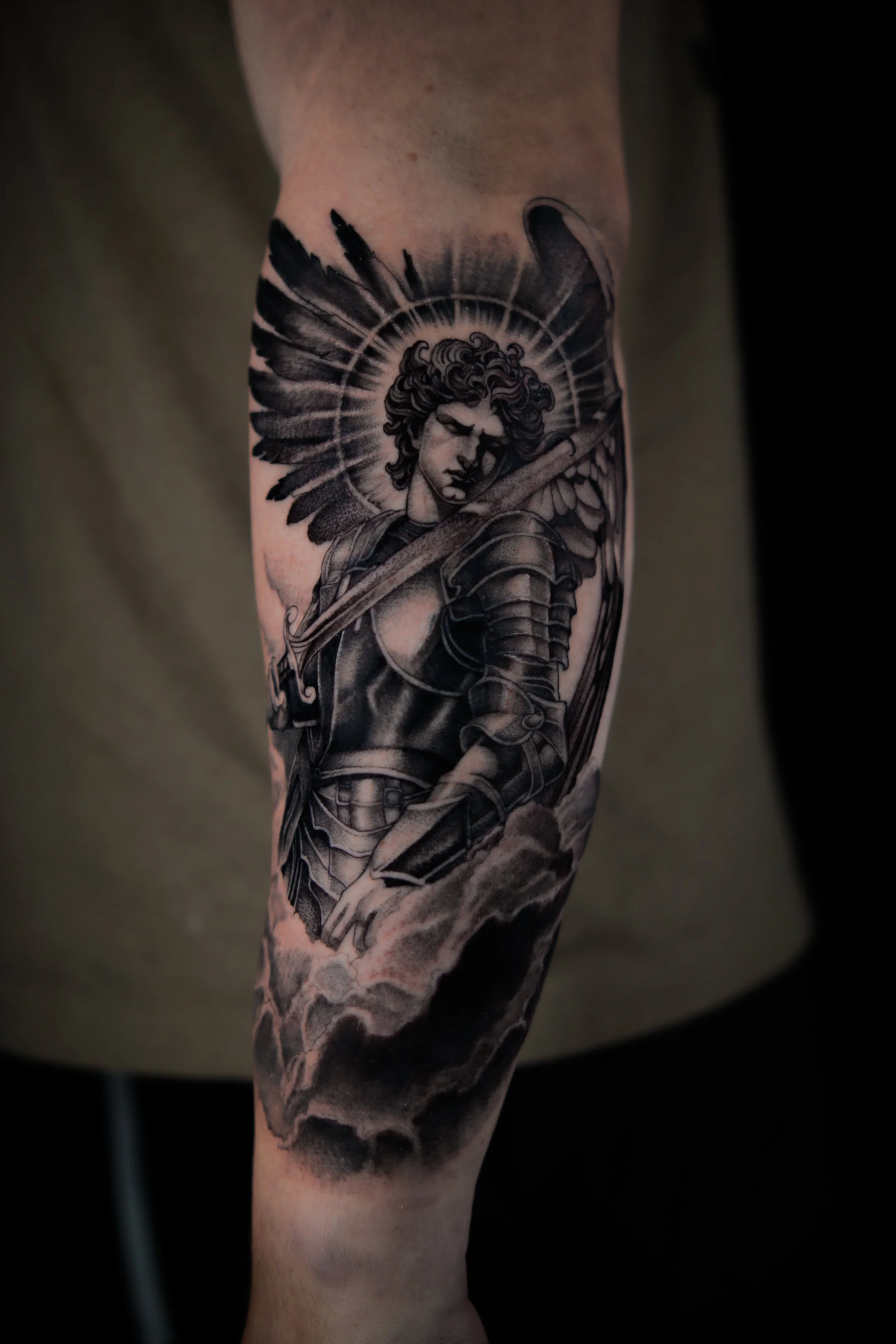 Black and gray tattoo of a warrior woman with wings, armor, and a sword, with a halo-like circle behind her head, and clouds below.