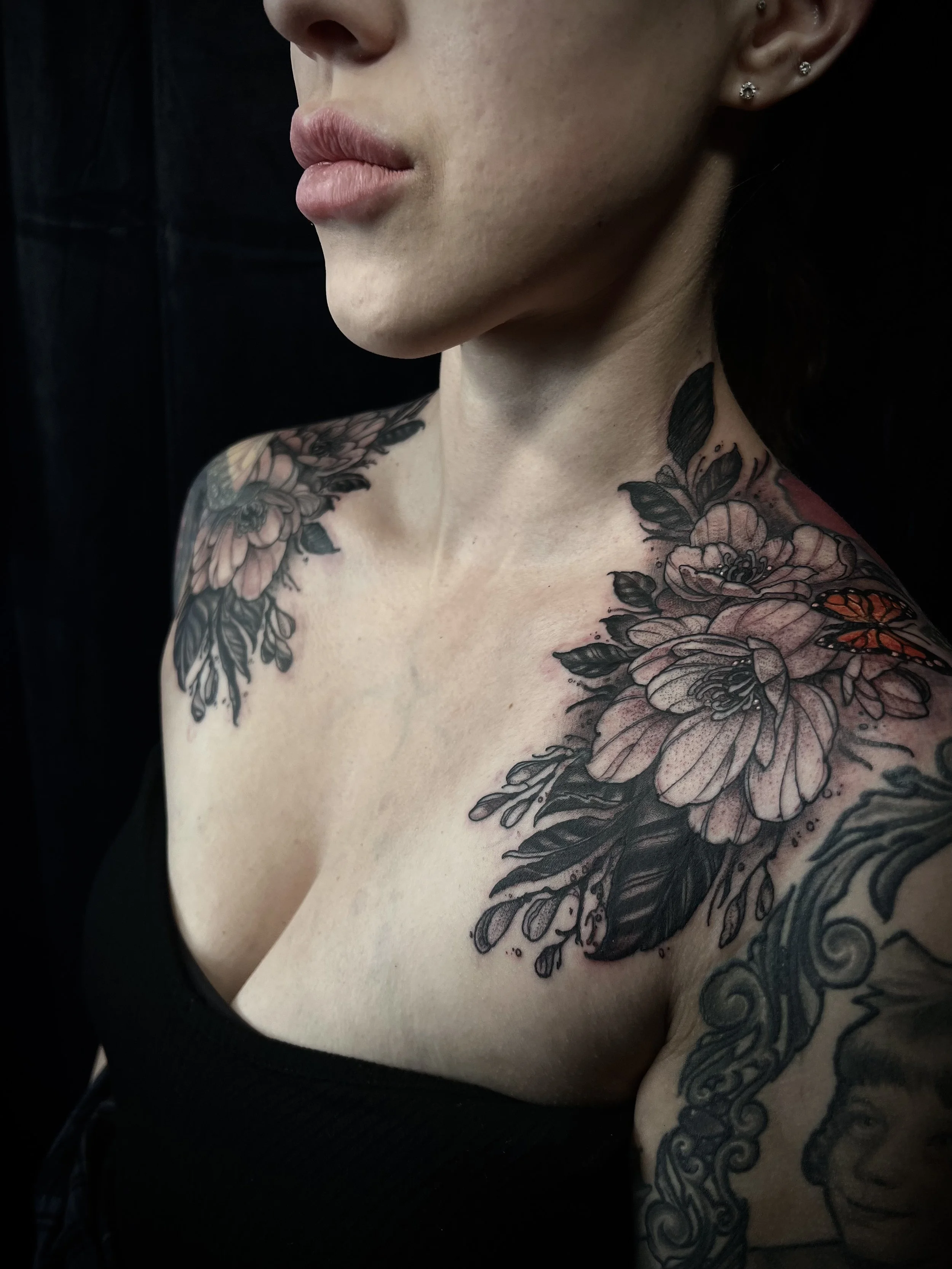 Close-up of a woman's shoulder tattoo featuring large pink flowers, black leaves, and a small orange butterfly, with additional tattoos visible on her arm.