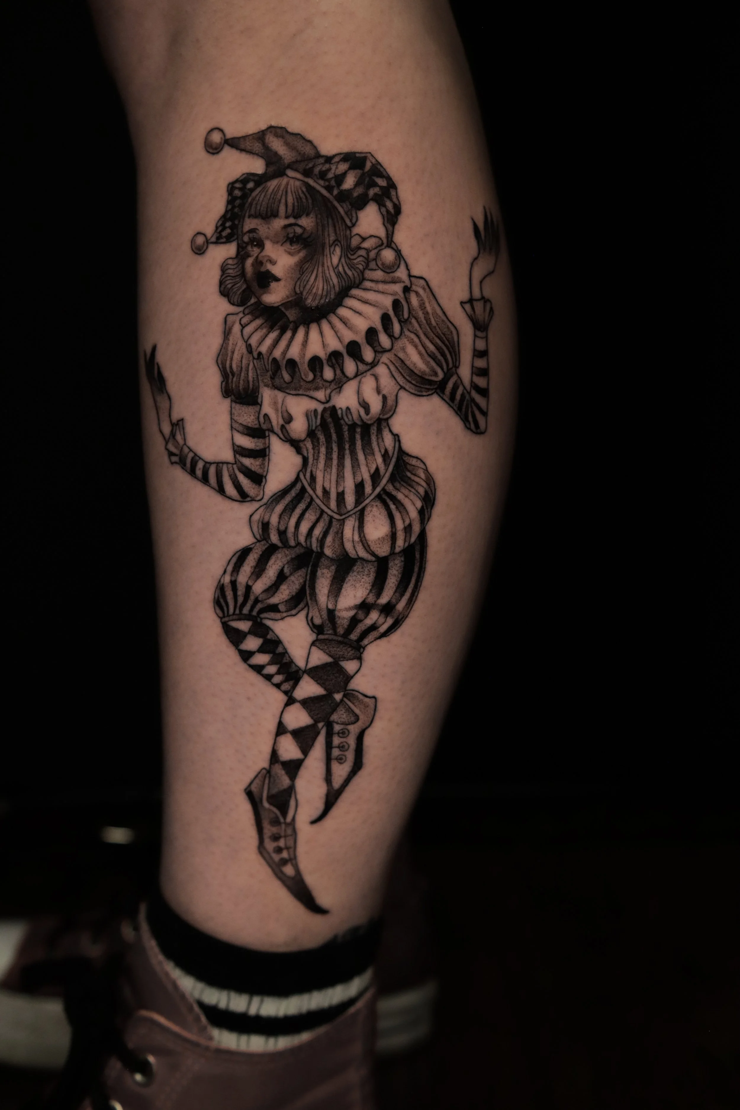 Black and gray tattoo of a harlequin clown on a person's leg. The clown has a female face with short hair, a ruffled collar, striped and checkered clothing, and pointed shoes with laces.